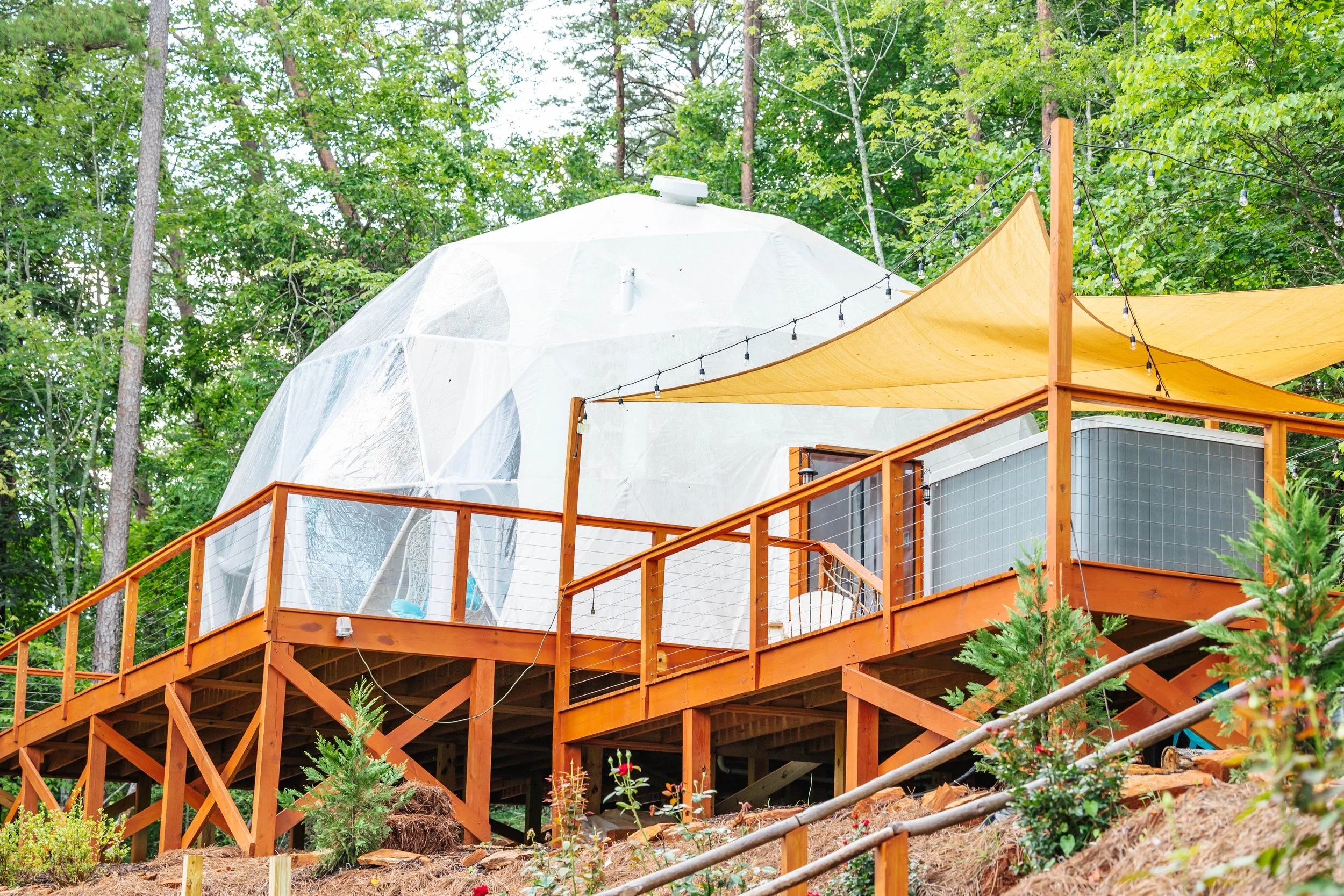 Treehouse with wooden deck, yellow shade sail, and transparent dome structure in a wooded area.