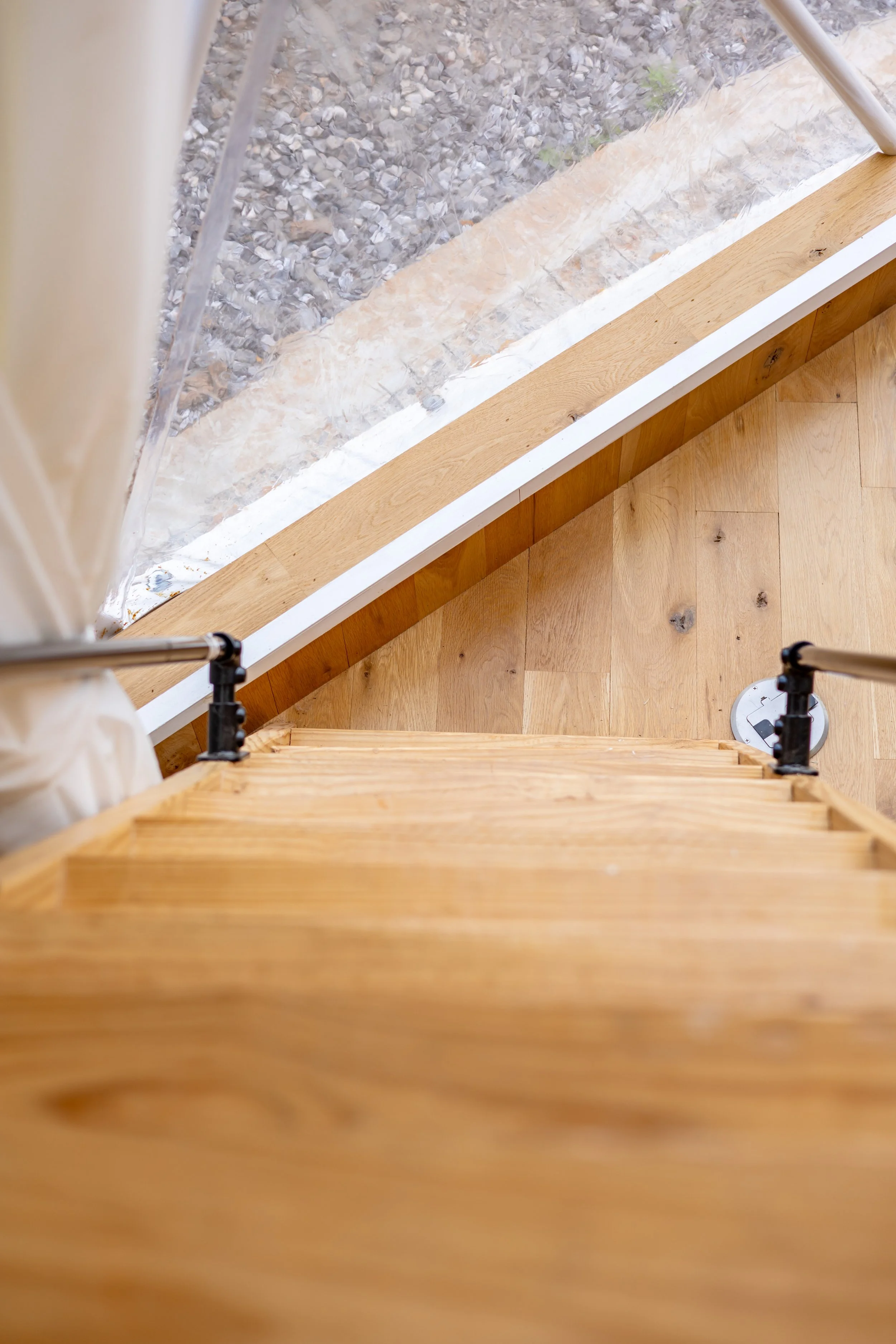 A view from the top of a wooden staircase looking down at a room with hardwood flooring, a white window frame, and outside gravel ground.