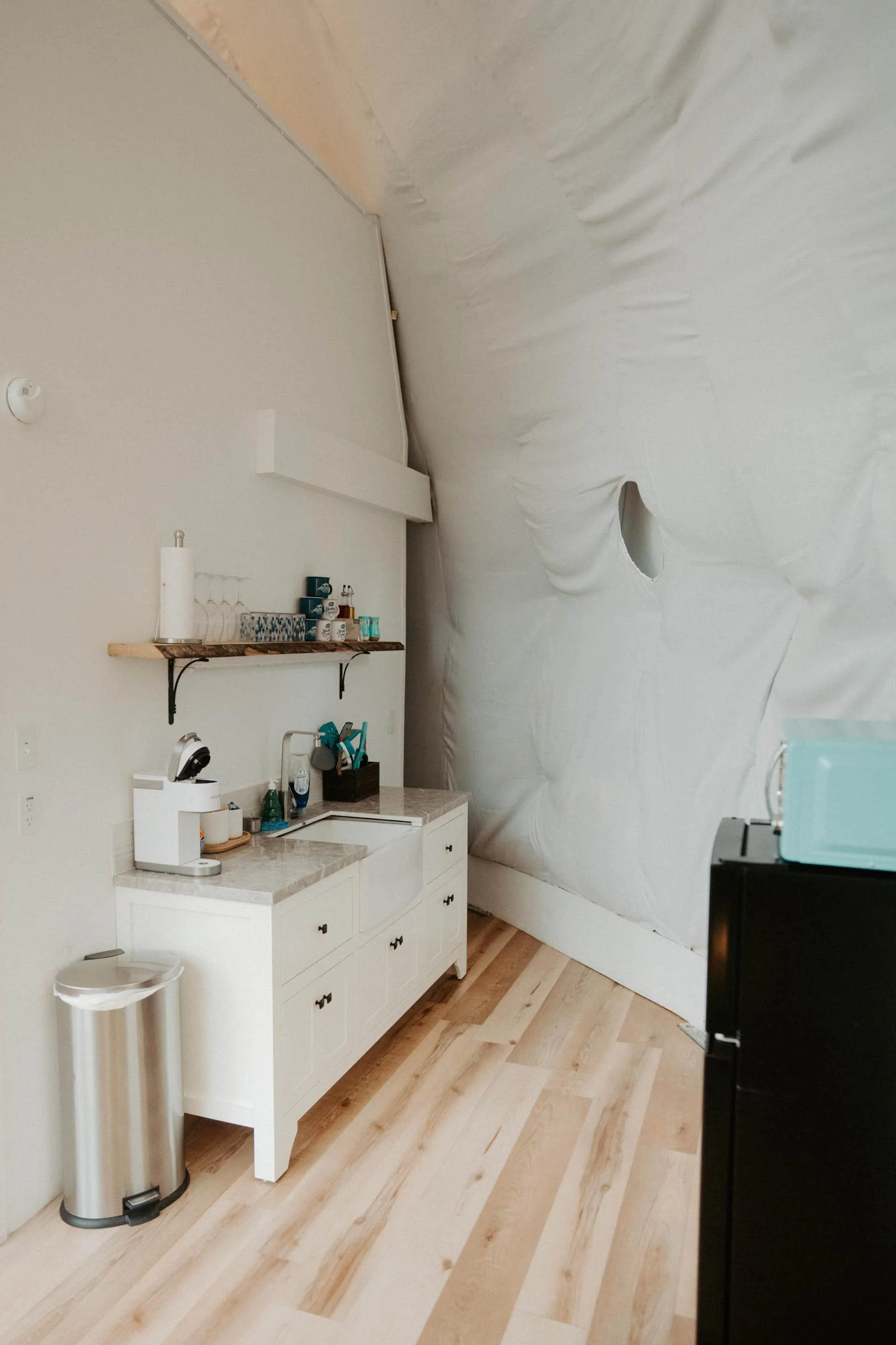 A small kitchen area with a white cabinet, a marble countertop, a black trash bin, and a black refrigerator. The wall behind is unfinished with insulation visible, and there is a wooden shelf holding glasses, cups, and small containers.
