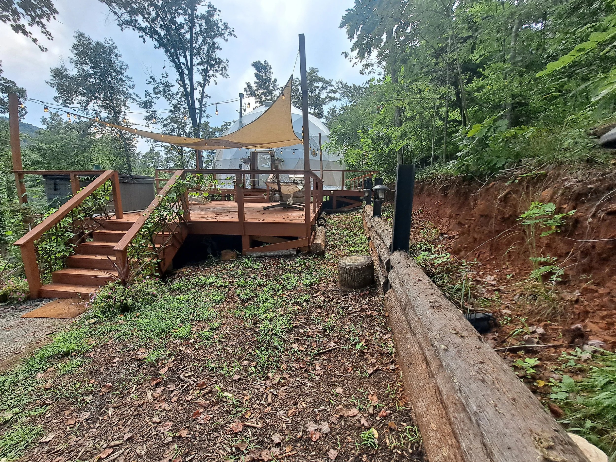 Wooden deck with small stairs leading up to it, surrounded by forested area with trees and greenery. There is a raised canopy or tent-like structure on the deck with string lights hanging above. A log is placed along a wooden railing, and a tree stum