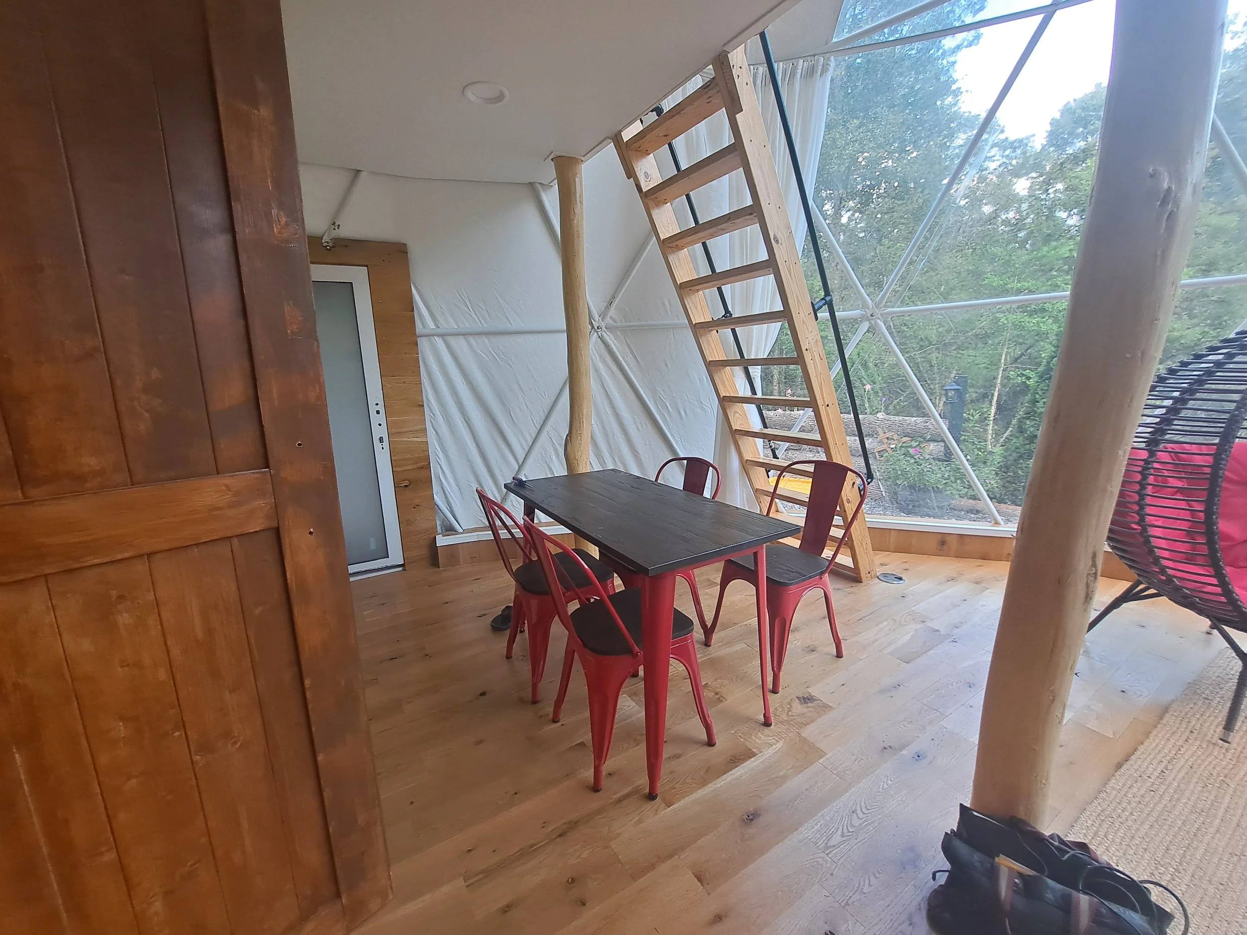 Interior of a modern treehouse with wooden floors, a black dining table, red chairs, a ladder leading to a loft, and large windows showing an outdoor view of trees.