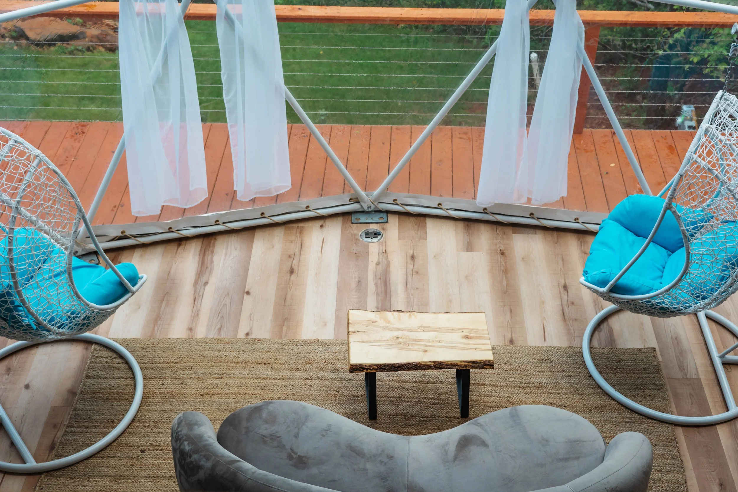 Seating area on a wooden deck with two hanging chairs with blue cushions, a small wooden table, a gray sofa, a rug, and white curtains overlooking a grassy yard.