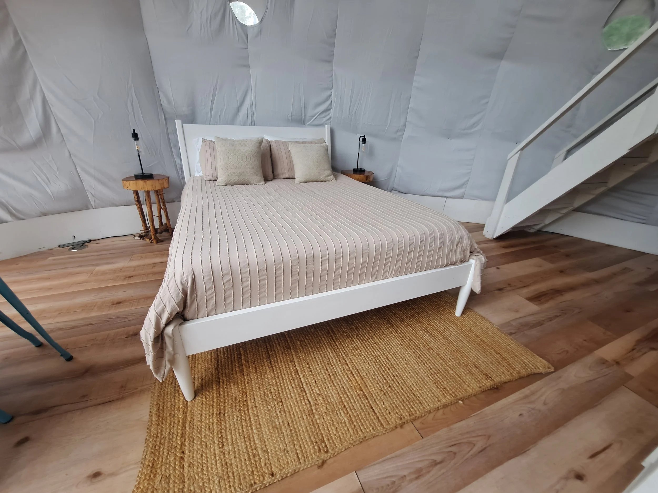 A bedroom with a white bed frame, cream striped bedding, and three pillows, a woven rug, two small wooden nightstands with black lamps, a staircase, and a wall covered with white fabric or paper.