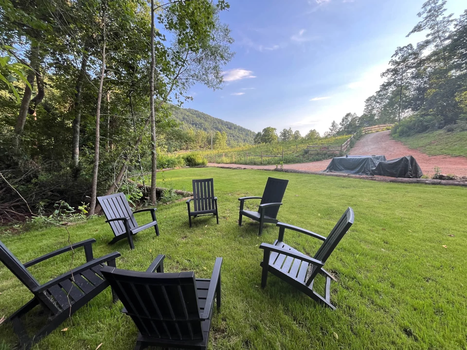 A circle of black Adirondack chairs on a grassy lawn next to a wooded area, with a dirt path and hill in the background under a clear blue sky.