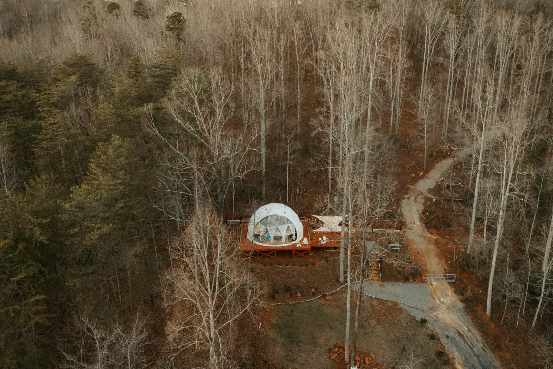 Aerial view of a dome-shaped glass house with a wooden deck in a forest, with leafless trees and winding dirt paths nearby.