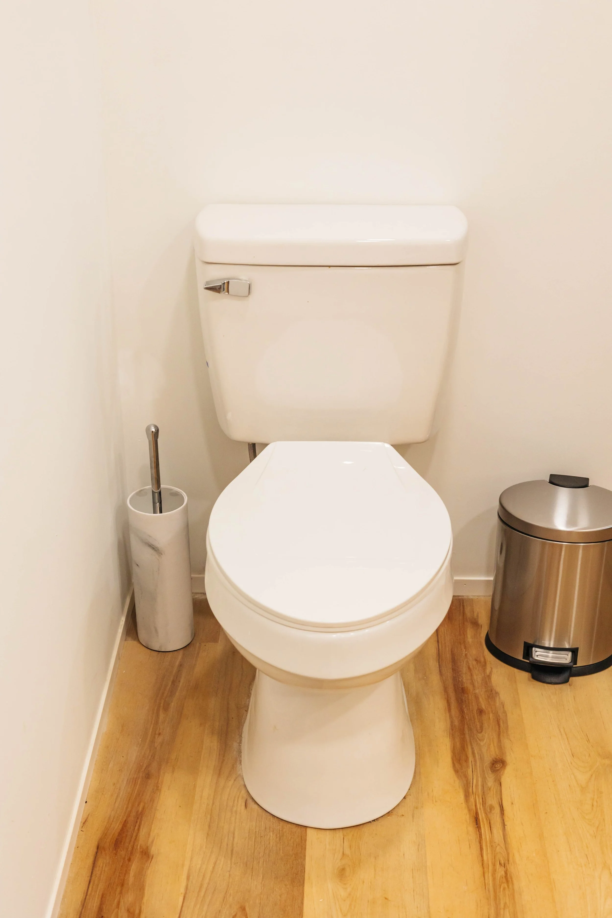 White toilet with a tank, toilet paper holder on left, and trash can on right, in a bathroom with wood flooring.