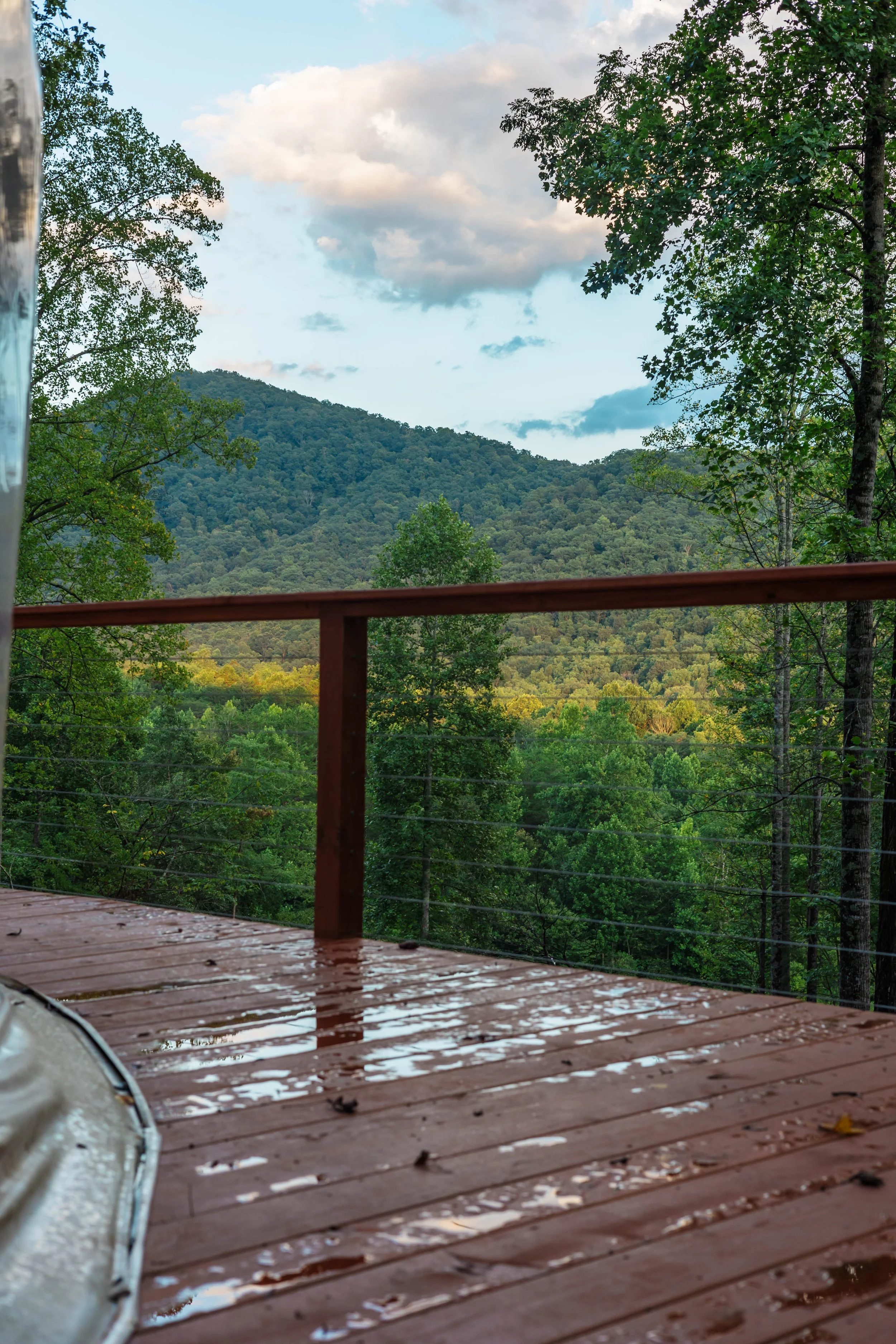 A wooden deck wet from rain with a view of green mountains and trees under a partly cloudy sky.