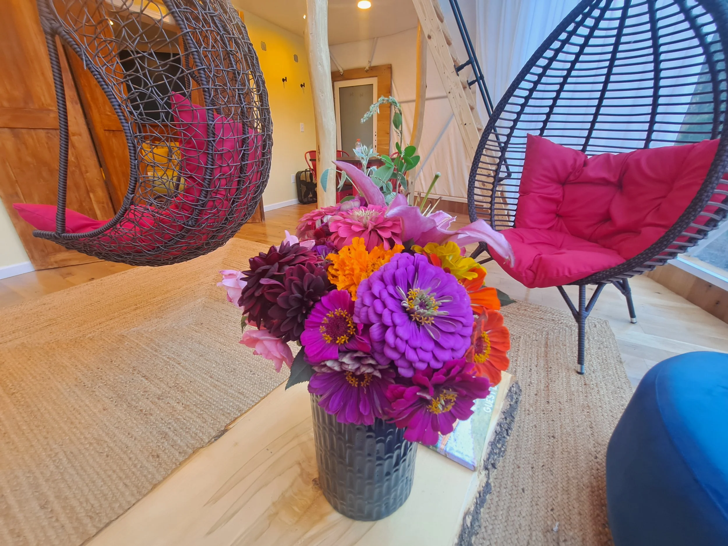 A bouquet of vibrant pink, purple, orange, and yellow flowers in a textured gray vase on a wooden coffee table in a cozy living room with hanging egg chairs and wooden walls.