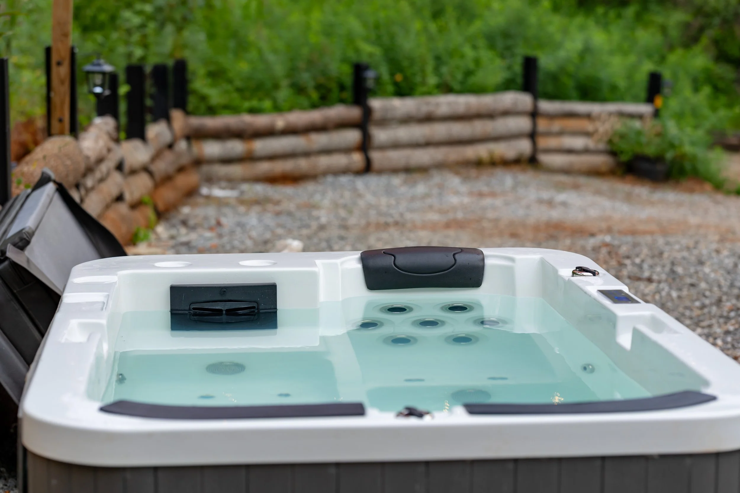 Outdoor hot tub filled with water, situated on gravel ground with a wooden fence and green trees in the background.