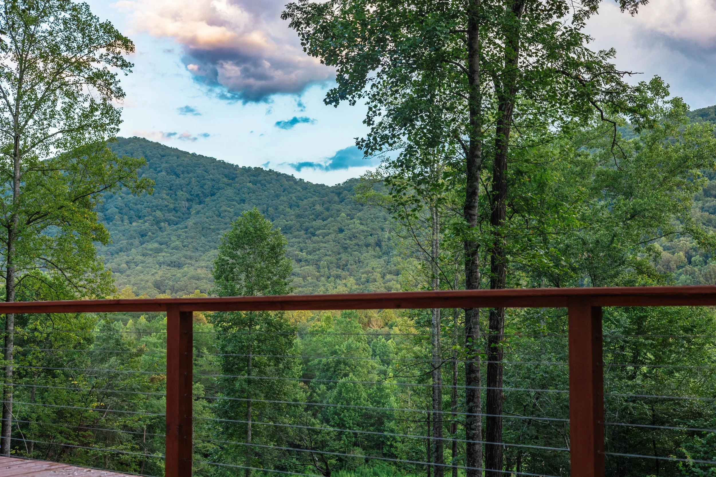 View of green trees and forested mountains under a partly cloudy sky, seen from a wooden deck with a railing.