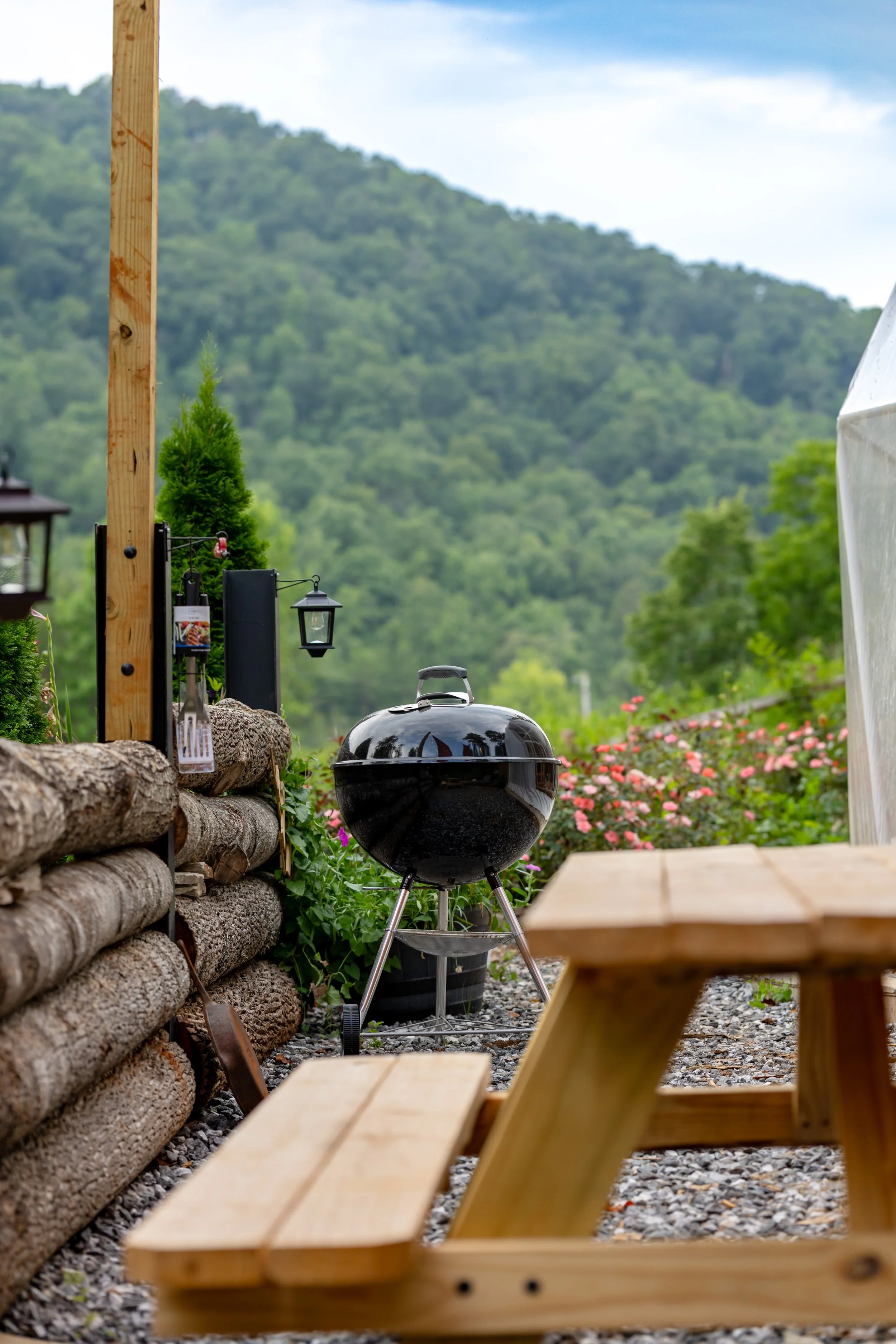 A black, round charcoal grill next to a wooden picnic table outdoors with flowers and trees in the background.
