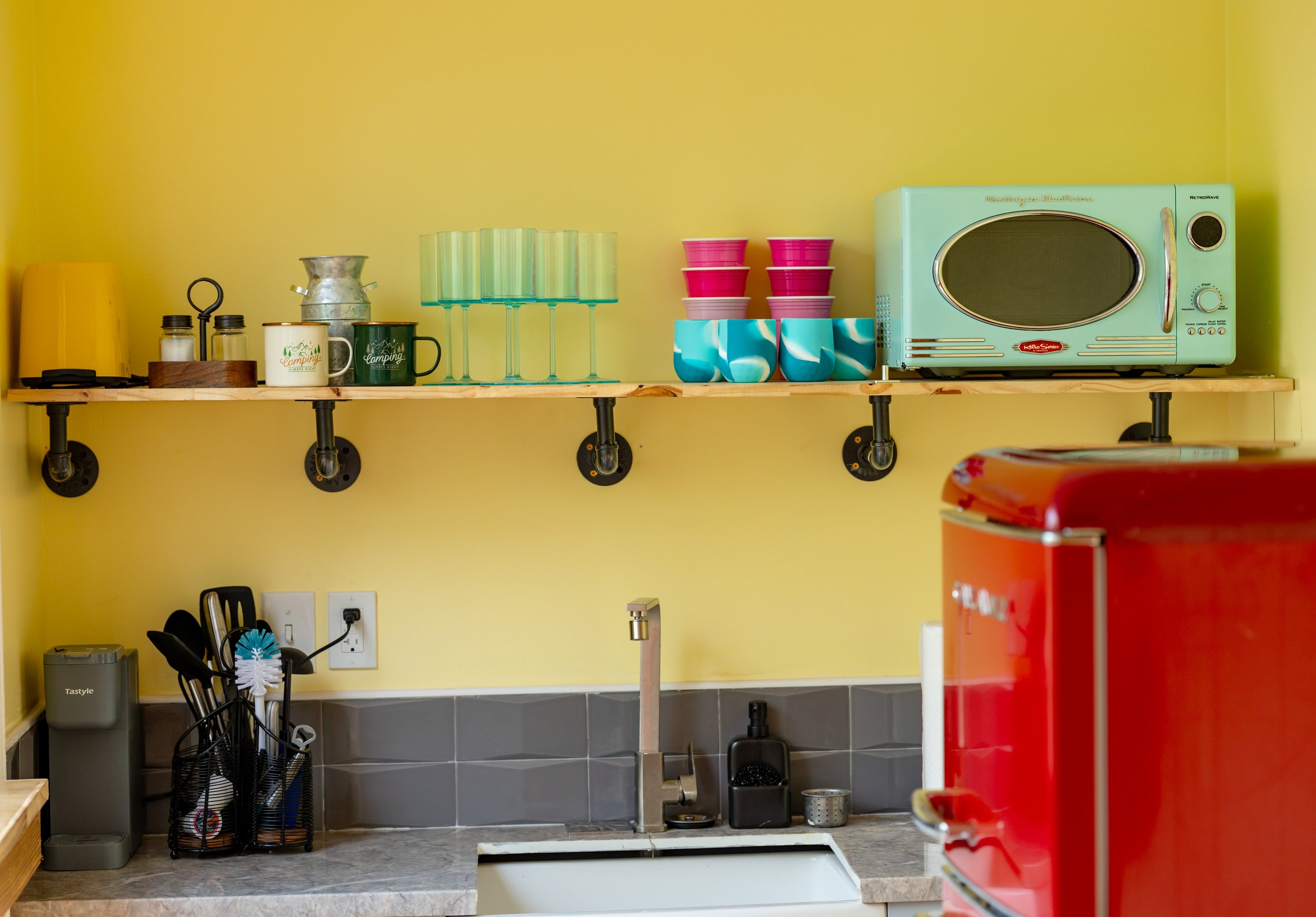 Kitchen shelf with a microwave oven, colorful cups, mugs, glasses, and storage jars on a yellow wall background.