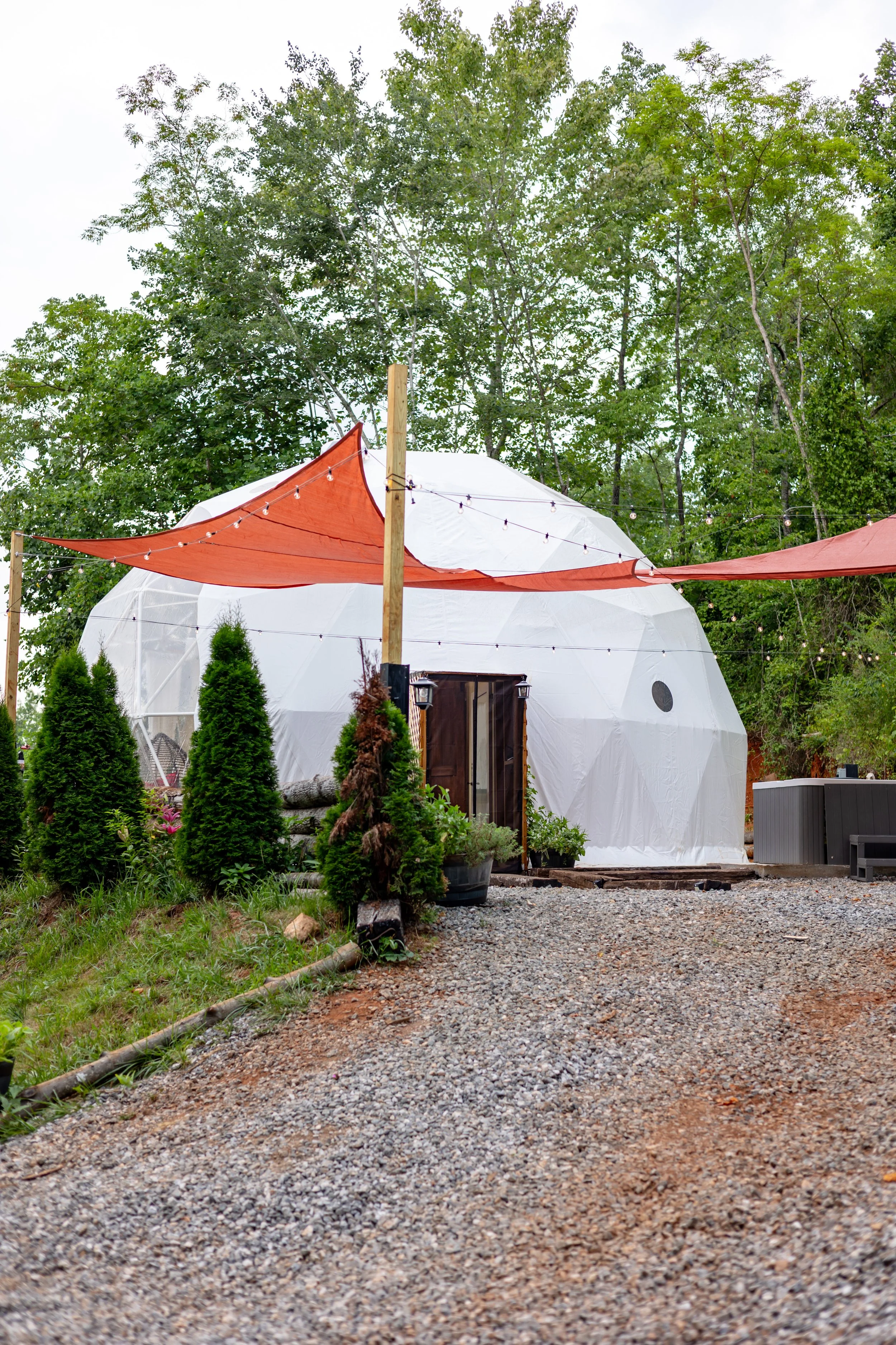 A geodesic dome structure with white covering, surrounded by greenery and string lights, with orange shade sails overhead and outdoor furniture nearby.