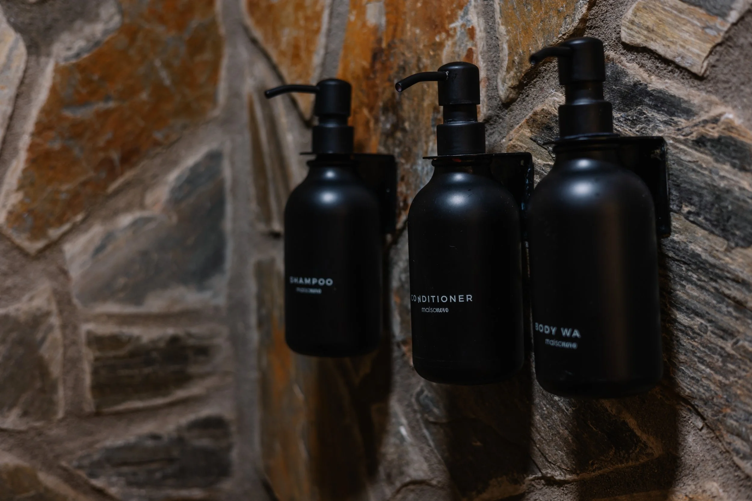 Three black bottles labeled shampoo, conditioner, and body wash mounted on a stone wall.