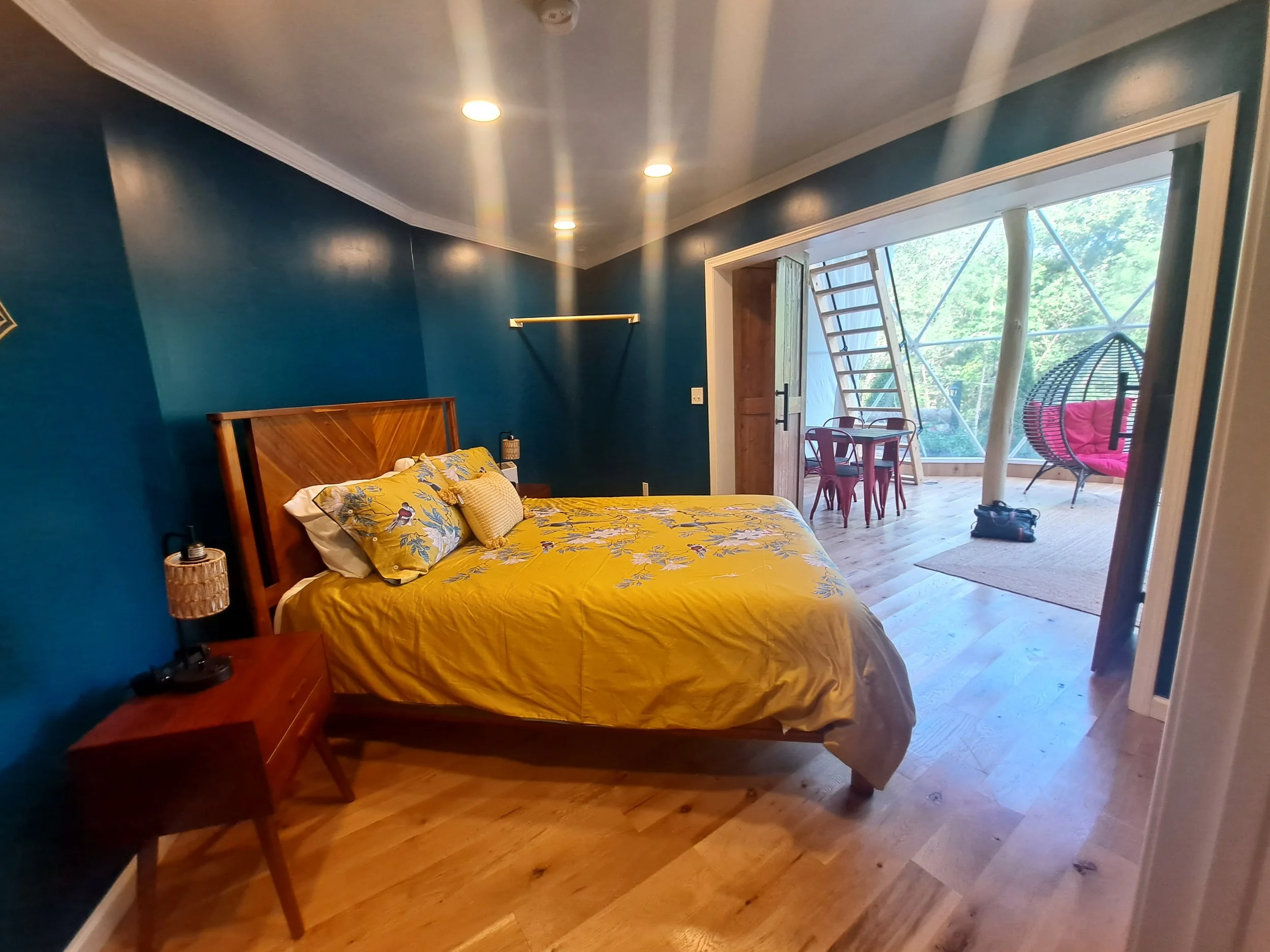 A bedroom with blue walls and wooden flooring. It has a wooden bed with yellow floral bedding and pillows. There are two bedside tables with lamps. An open door leads to a sunlit enclosed porch with a table, chairs, a hanging chair with pink cushions