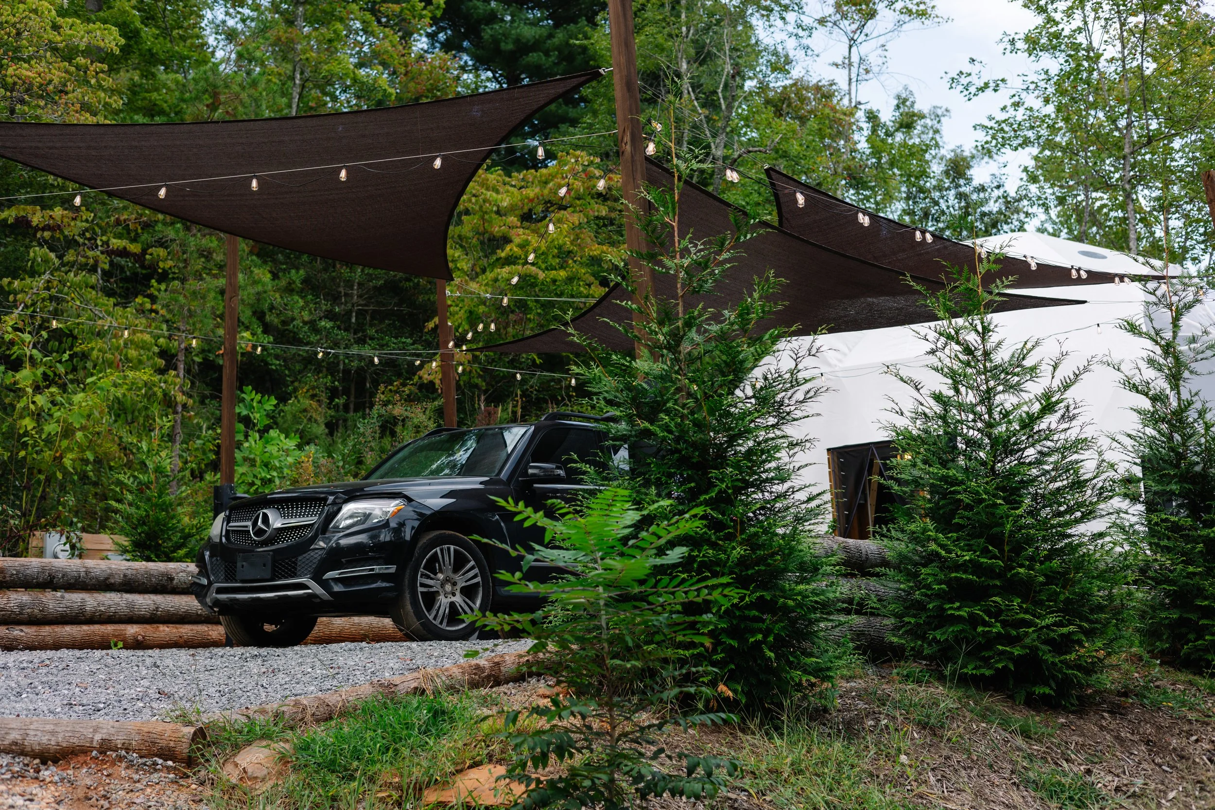 A black Mercedes-Benz SUV parked next to a white tent structure on a gravel area, surrounded by green trees and shrubs, with brown shade sails and string lights overhead.
