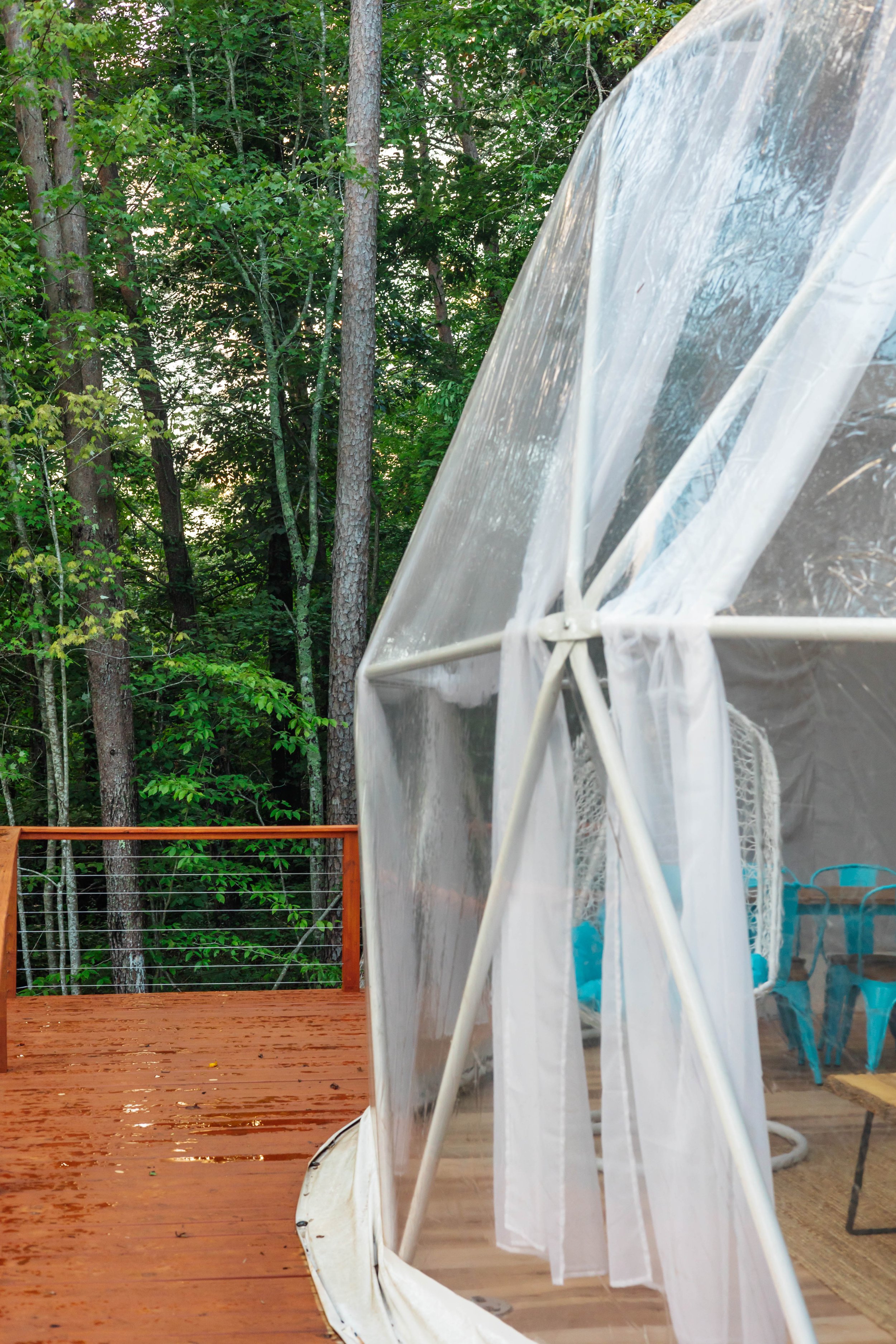 A wooden deck with rain droplets, beside a white garden tent with sheer curtains, overlooking a lush green forest.