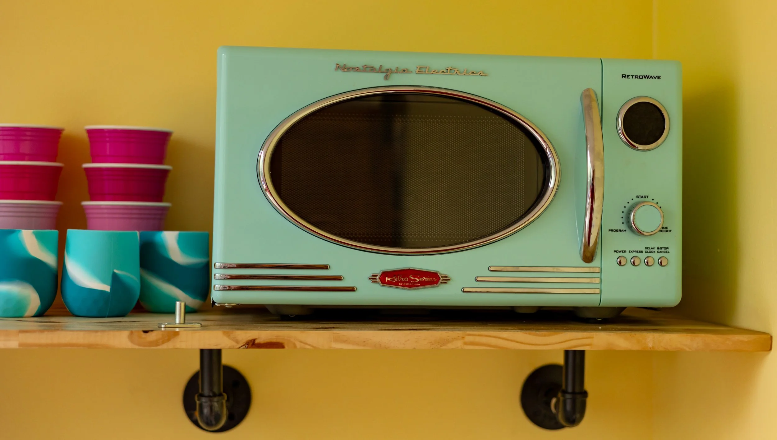 A vintage turquoise microwave oven on a wooden shelf against a yellow wall, next to pink bowls and turquoise cups.