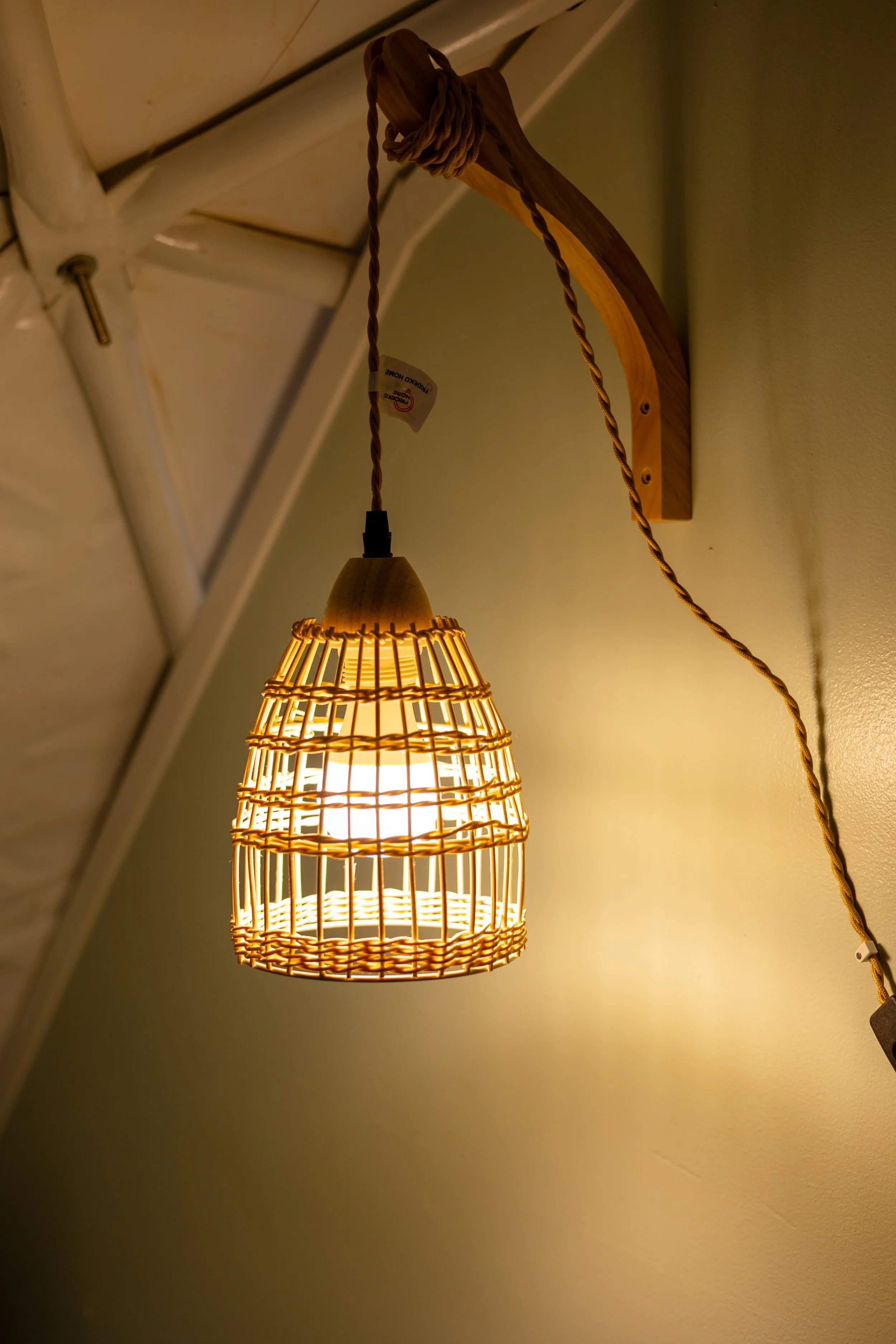 A hanging ceiling lamp with a basket-like design made of wooden and rattan materials. It is attached to a wall with a wooden bracket and features a twisted fabric cord.