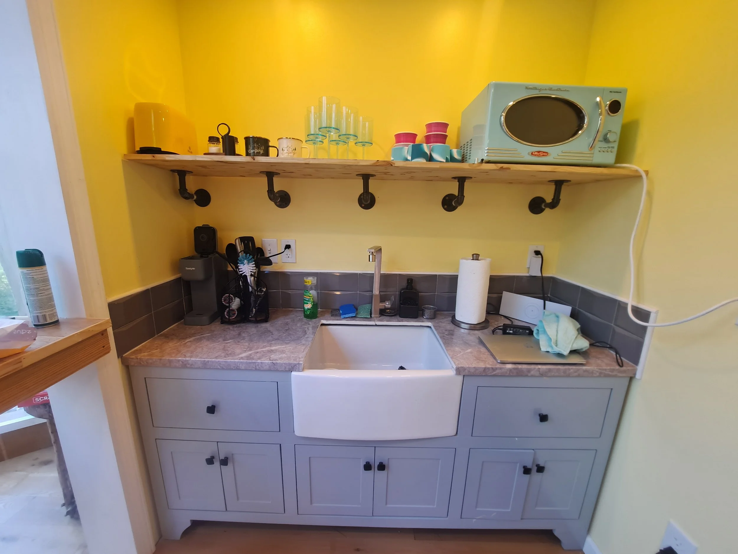 Kitchen sink area with yellow wall, open shelf holding glasses, mugs, and a microwave, gray cabinetry, and various small appliances and utensils on the countertop.