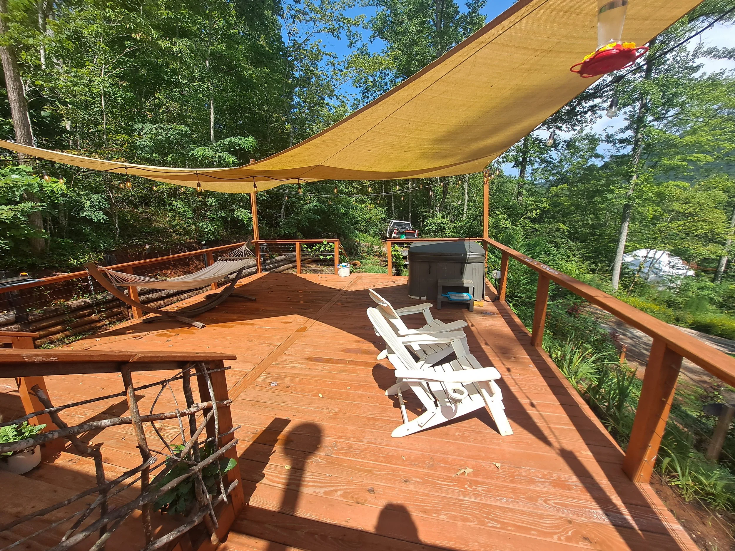 A wooden deck in a wooded area with two white patio chairs, a large hot tub, a hammock, and a beige canopy providing shade, surrounded by green trees.