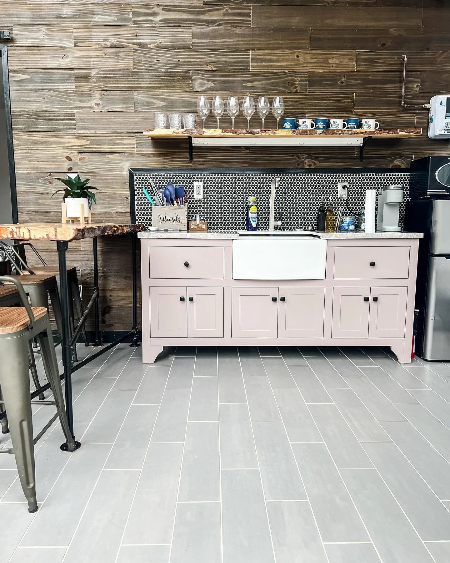 A kitchenette with a pink cabinet, a white farmhouse sink, and a wooden shelf holding wine glasses and coffee mugs against a wood-paneled wall.
