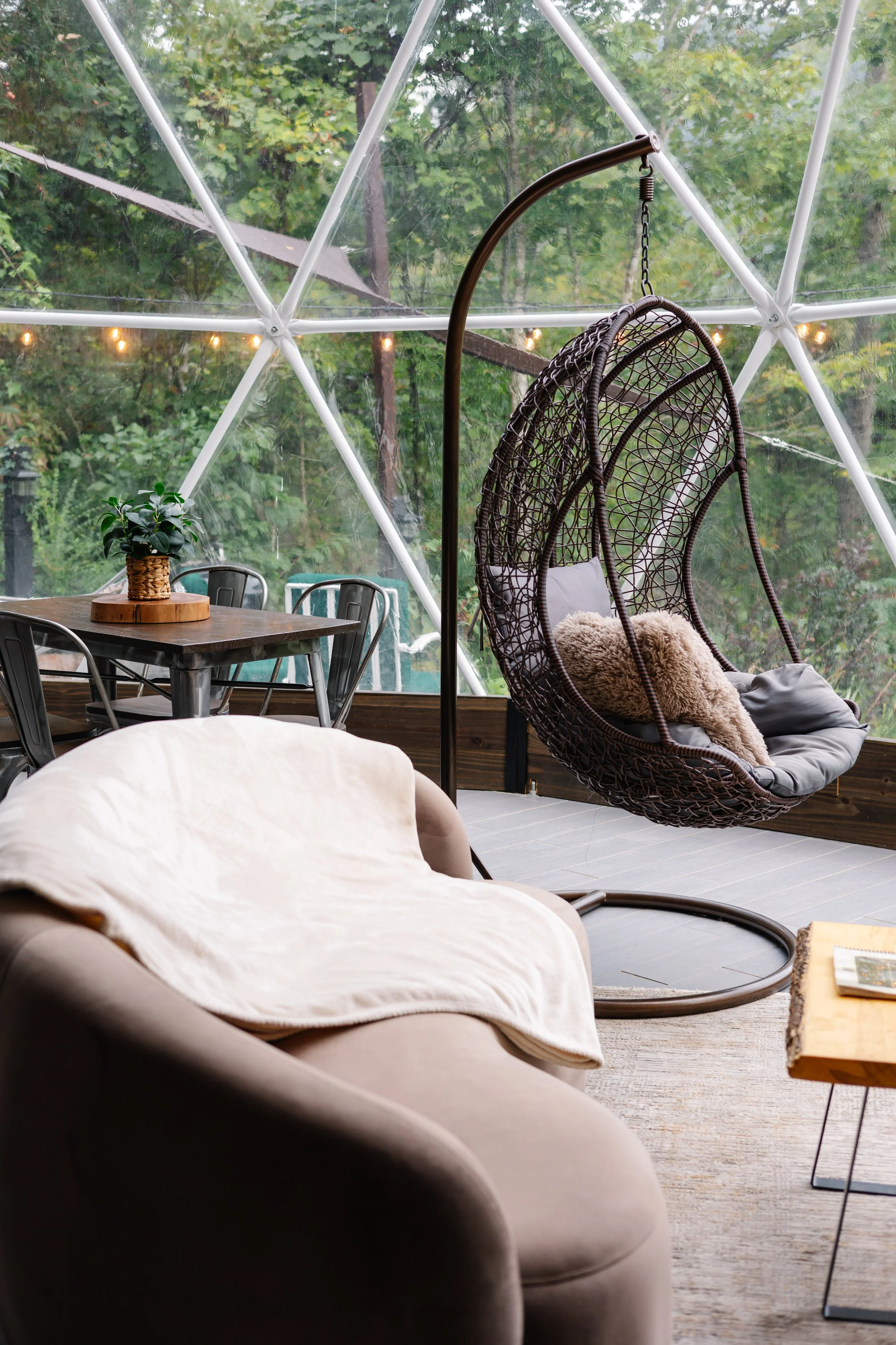 Living room with a beige sofa draped with a cream blanket, a wooden side table with a book, a black metal dining table and chairs, and a hanging wicker egg chair with cushions, all inside a geodesic dome with large windows showing green trees outside