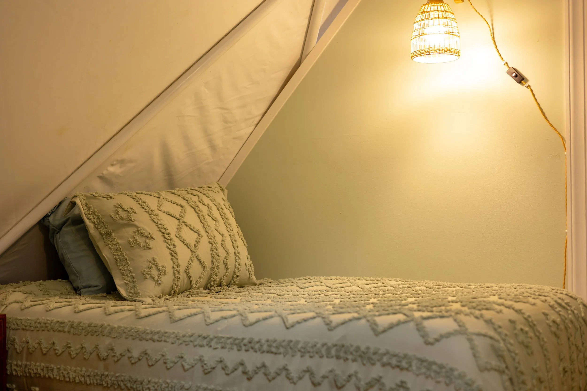 A neatly made bed with a textured, beige bedspread and matching pillow in a cozy bedroom. A wicker lampshade hangs on the wall, illuminating the room softly.