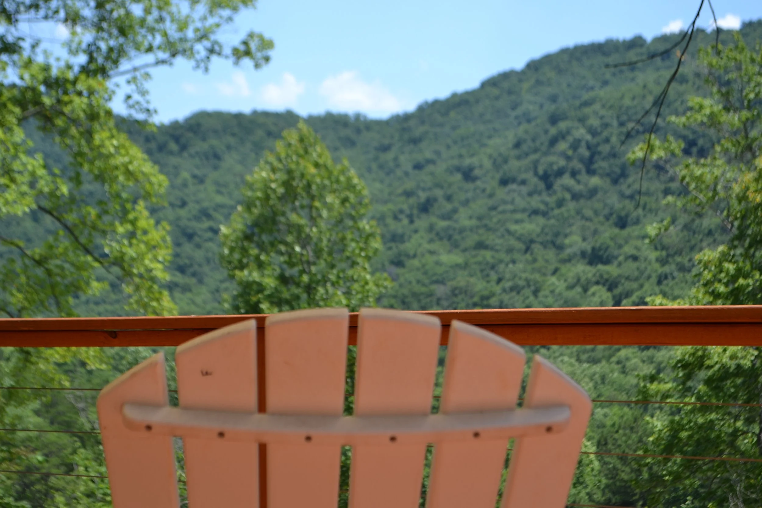 Pink Adirondack chair on a wooden deck overlooking green trees and mountains under a blue sky.