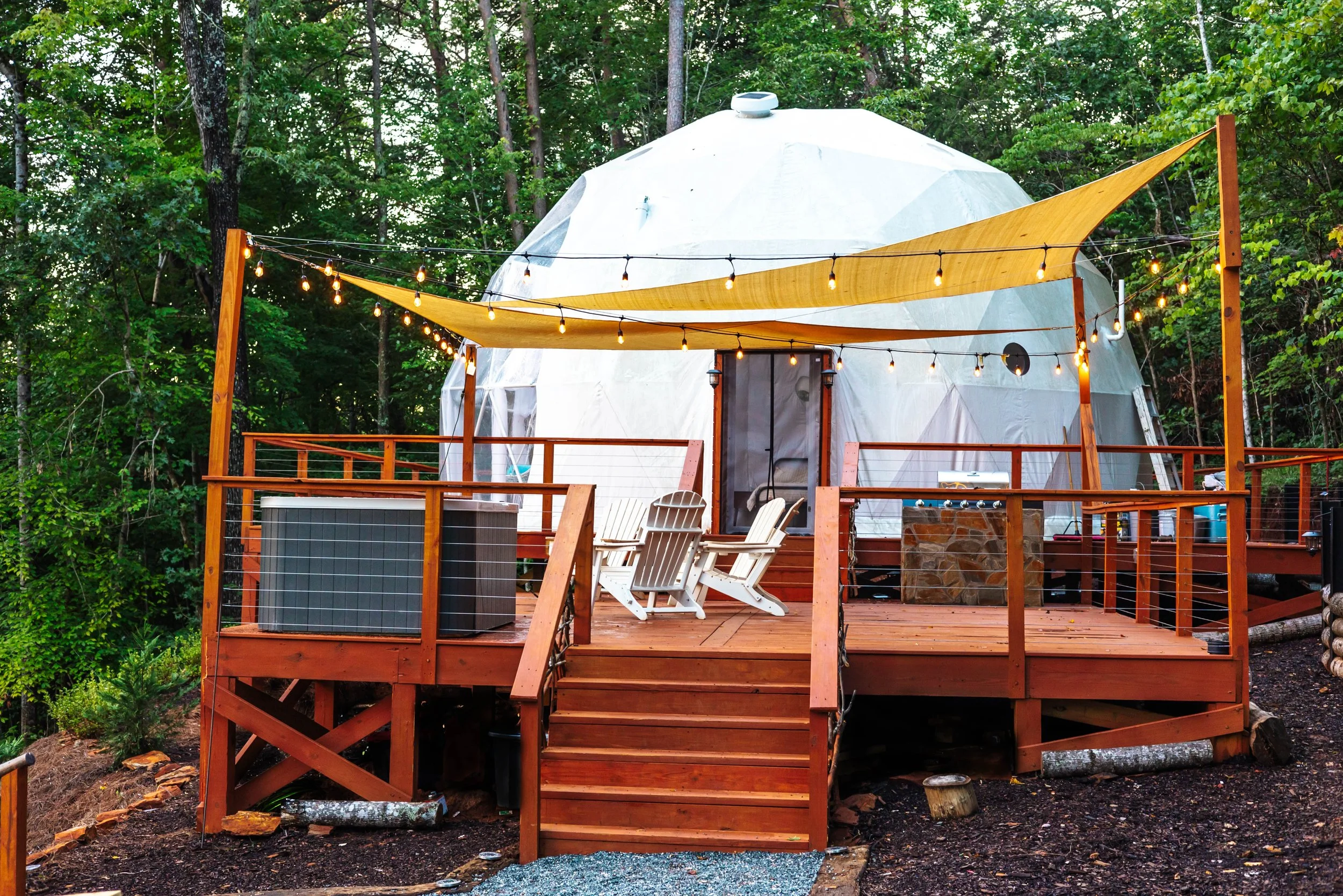 A large, dome-shaped glamping tent on a wooden deck, surrounded by trees. The deck has string lights, Adirondack chairs, and a small outdoor kitchen with a hot tub nearby.