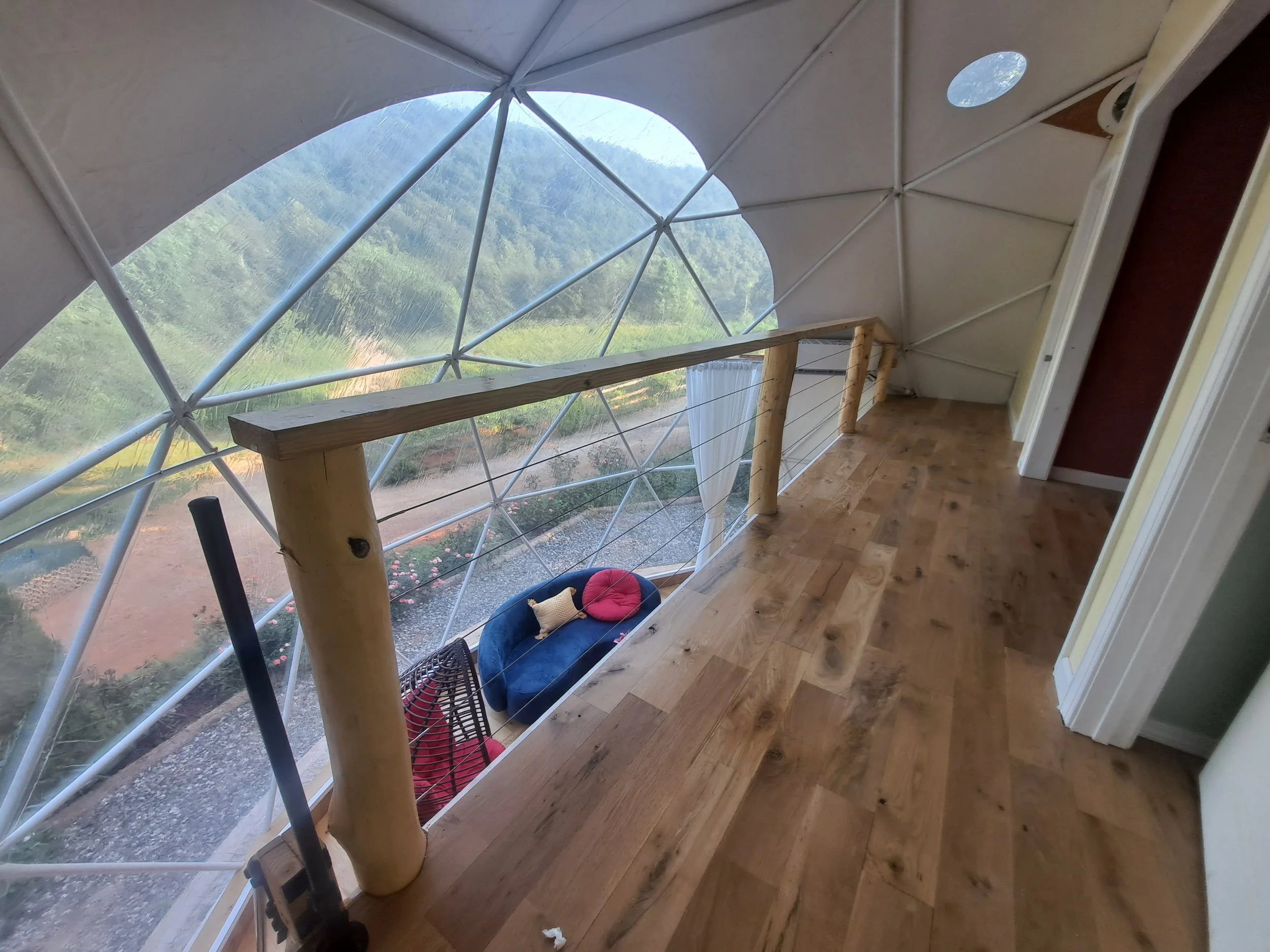 Interior view of a small room with a sloped ceiling and a large geodesic window overlooking a green mountainous landscape, wooden flooring, and a rustic wooden railing.