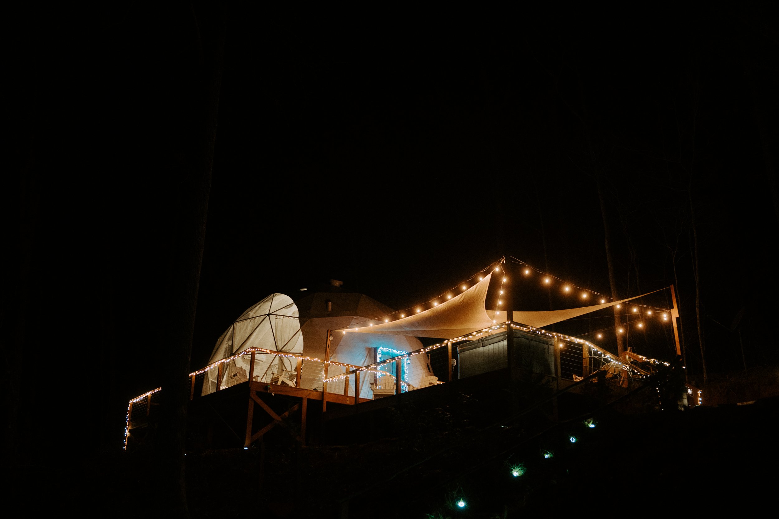 A nighttime outdoor scene featuring a deck with string lights and a covered area, possibly a dome or tent, illuminated against the dark sky.