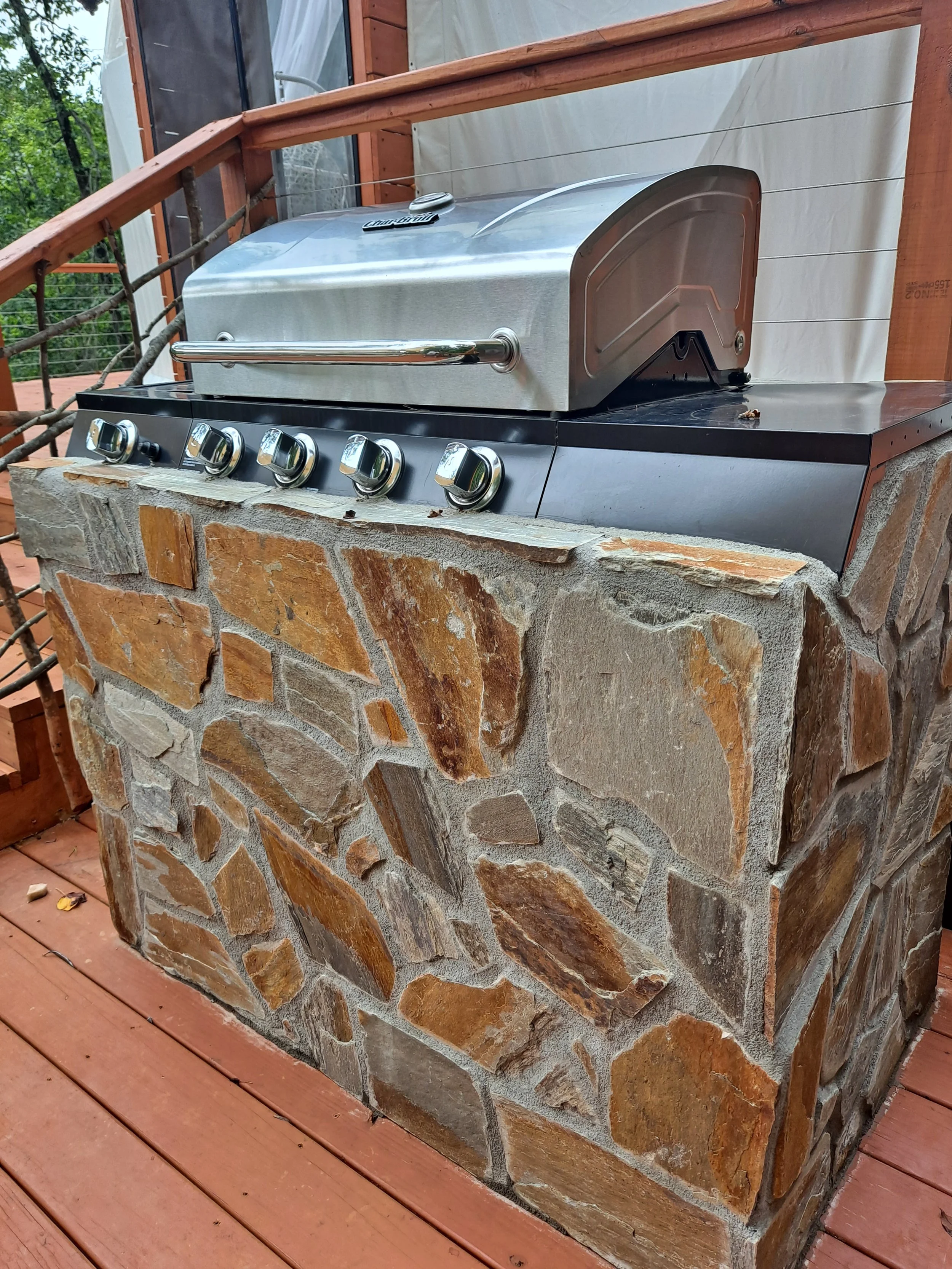 A stainless steel outdoor gas grill with five control knobs is built into a stone pillar on a wooden deck. The grill has a lid and a handle.