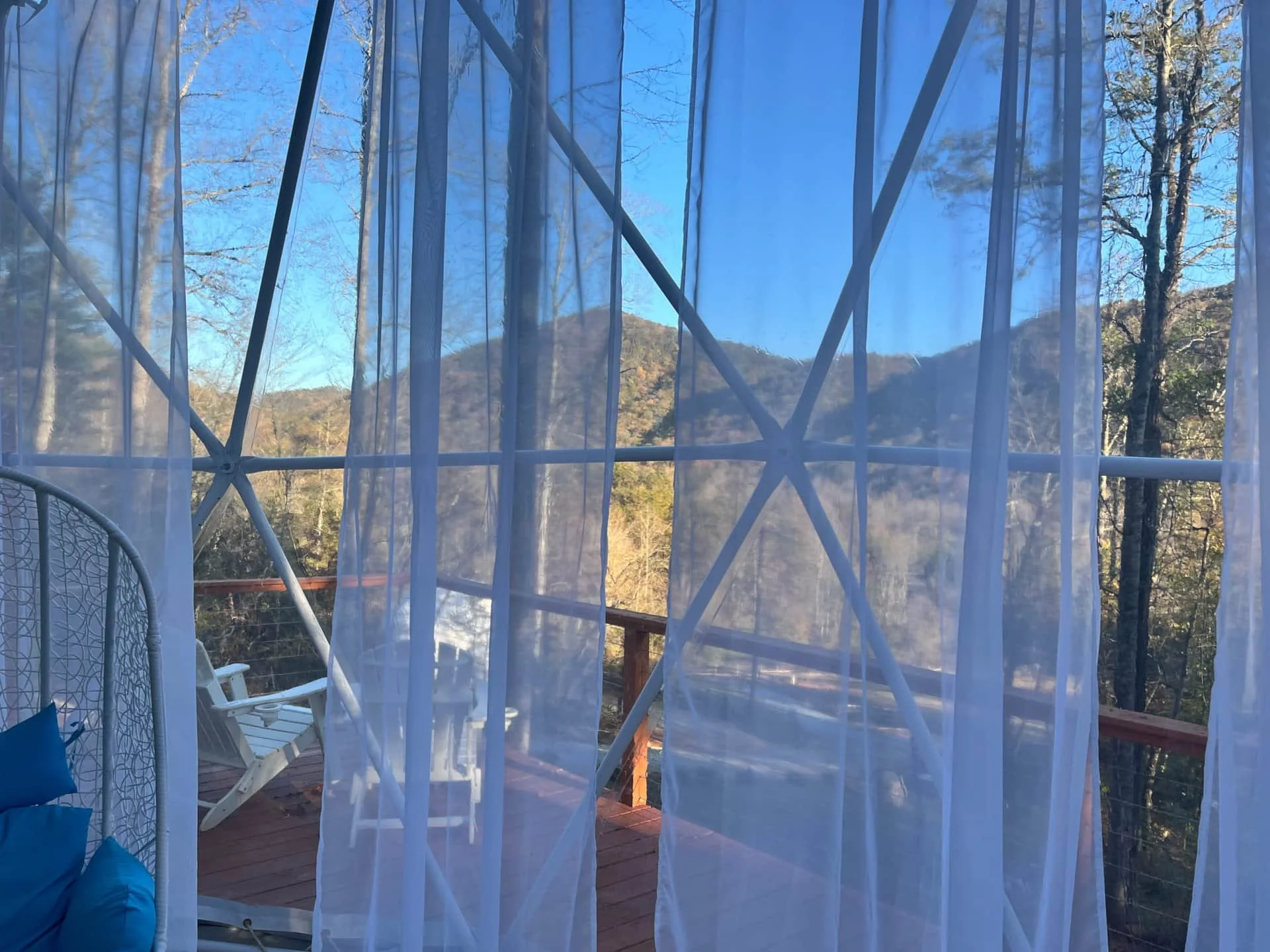 View through sheer white curtains overlooking a wooden deck with white chairs and a view of a mountain and clear blue sky.