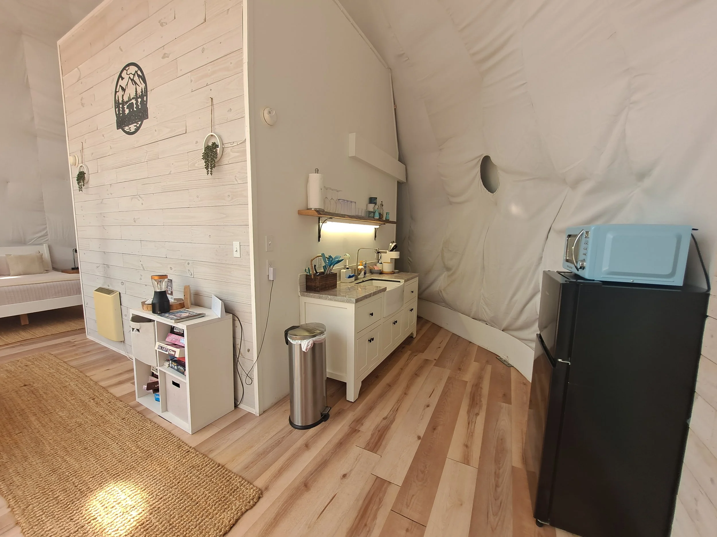 Interior of a cozy cabin with a small kitchenette, a bed visible in the background, and various kitchen appliances including a microwave and mini fridge, with wooden flooring and a light-colored wall.