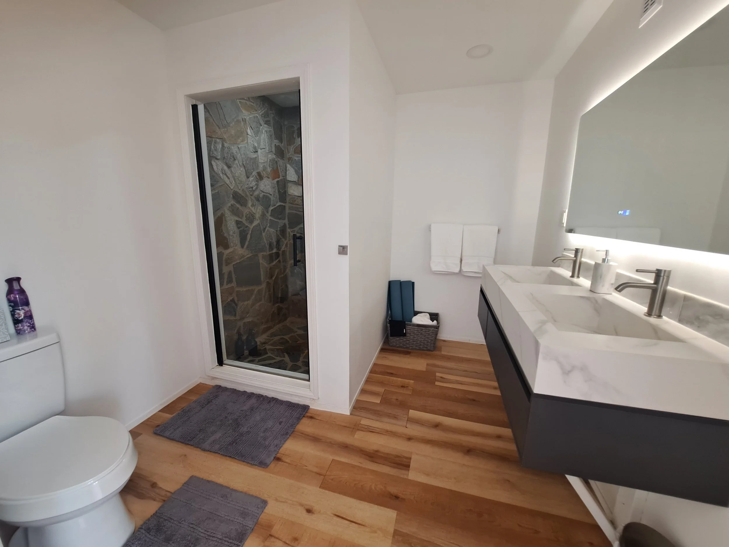 Modern bathroom with wooden floor, white walls, and a double sink marble vanity. Features a walk-in shower with a glass door and stone tile interior, a toilet with a purple bottle on top, and a basket with towels and mats on the floor.