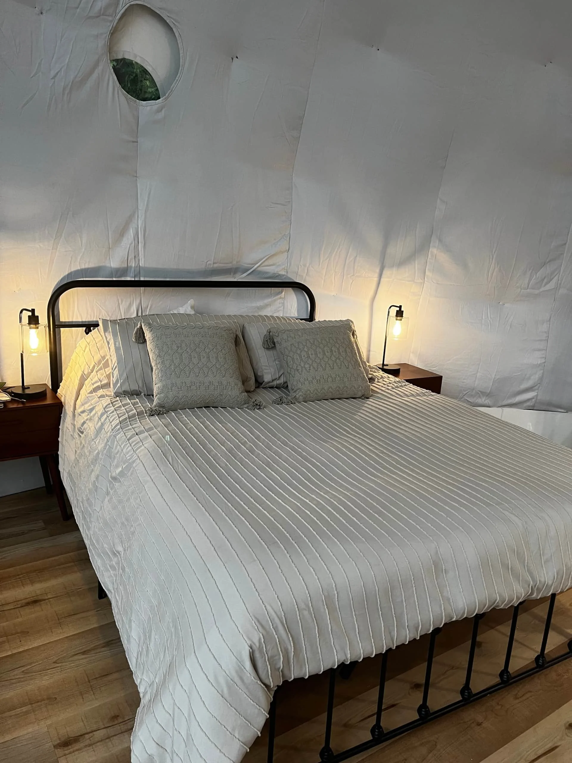 A neatly made bed with striped bedding, three decorative pillows, two small side tables with lamps, and a white textured wall with a circular window.