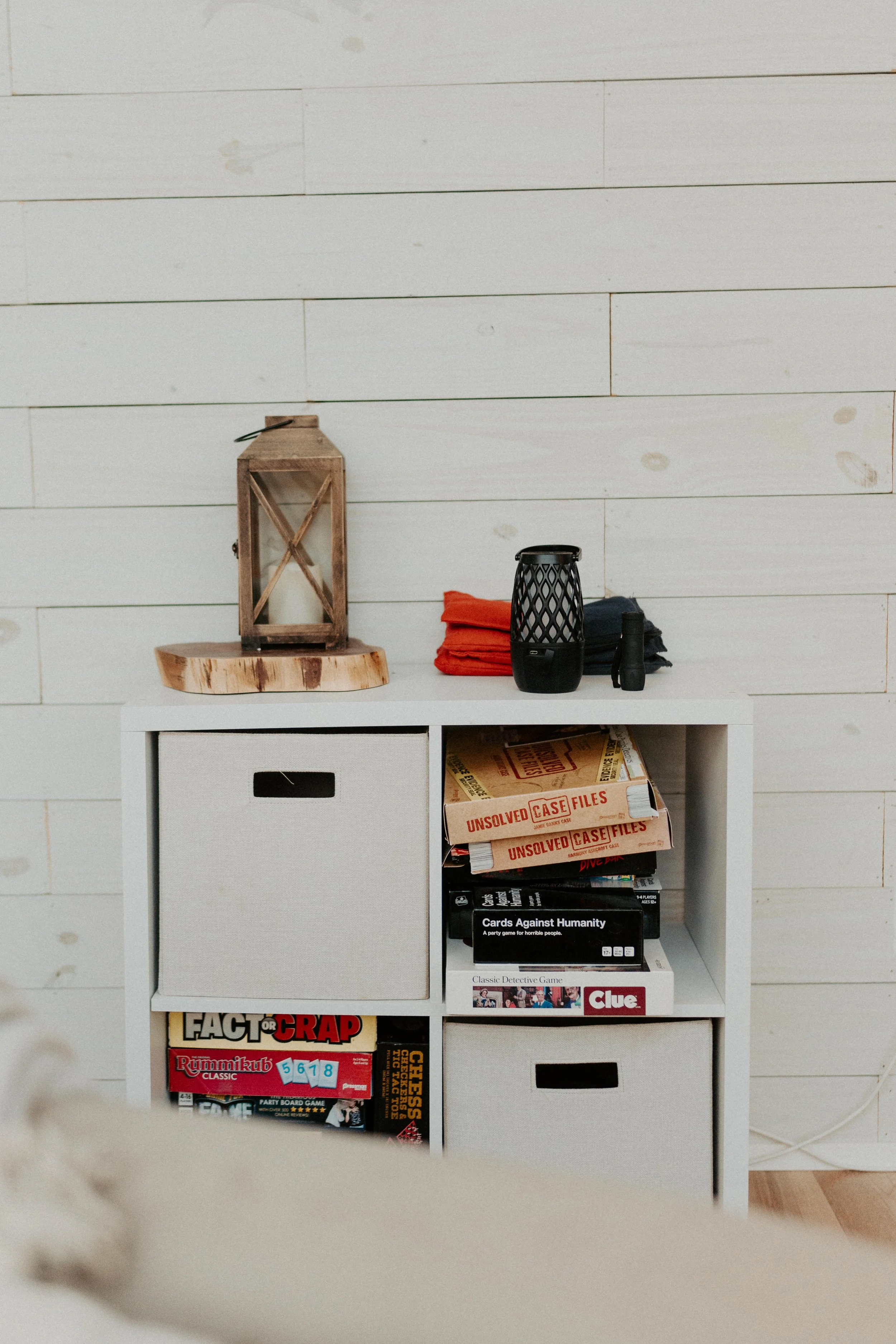 White cubby shelf with four compartments, some containing board games like "Fact or Crap," "Rummikub," and "Clue." On top of the shelf are a wooden lantern, folded red and black cloths, a black lattice lantern, and a small black object. The backgroun