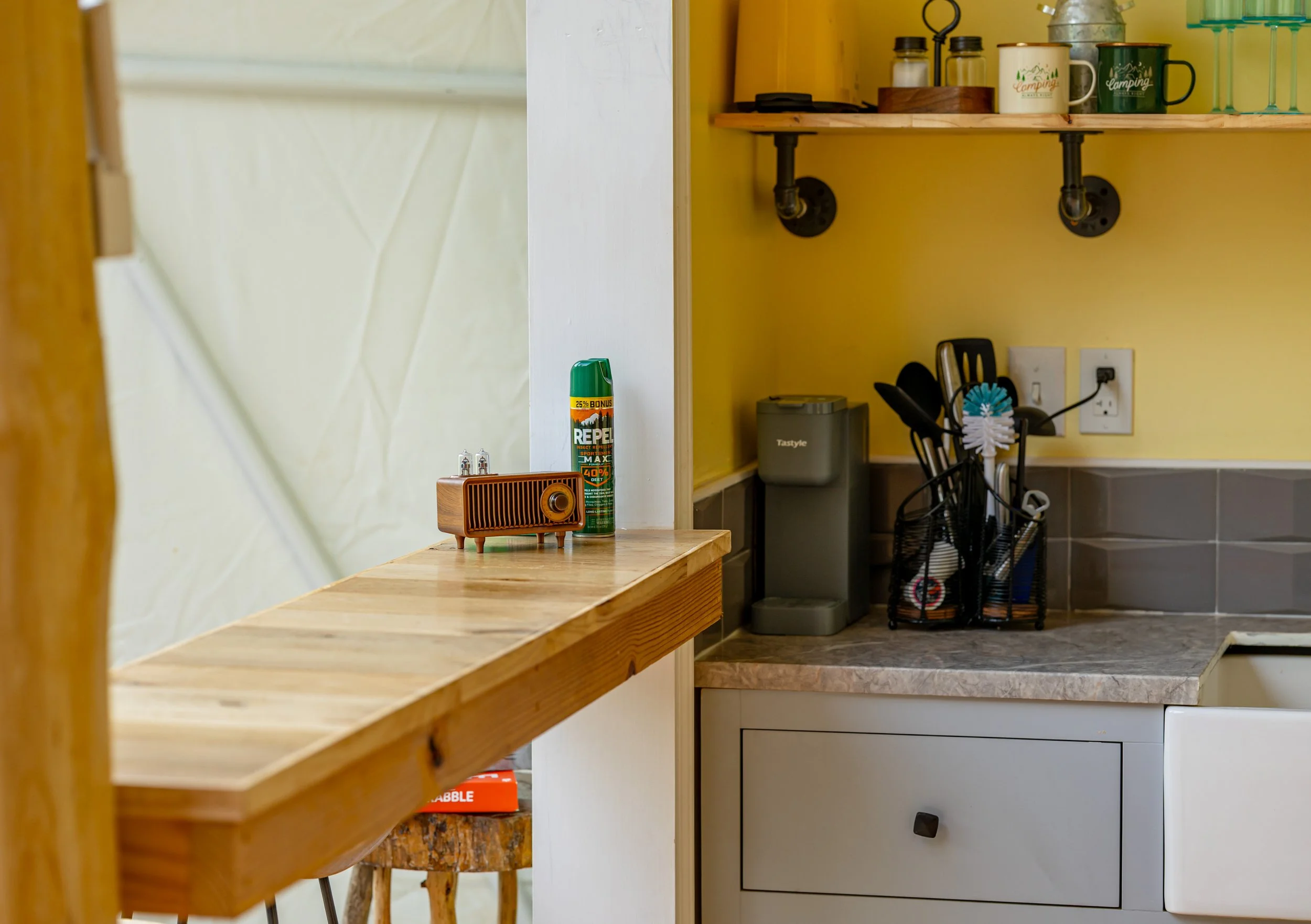 A kitchen counter with shelves, a coffee machine, utensils in a holder, and a box of coffee filters, with a yellow wall in the background.