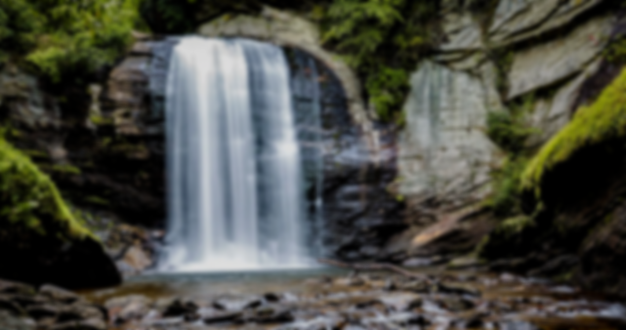 Blurry image of a waterfall cascading down rocks surrounded by lush green forest.