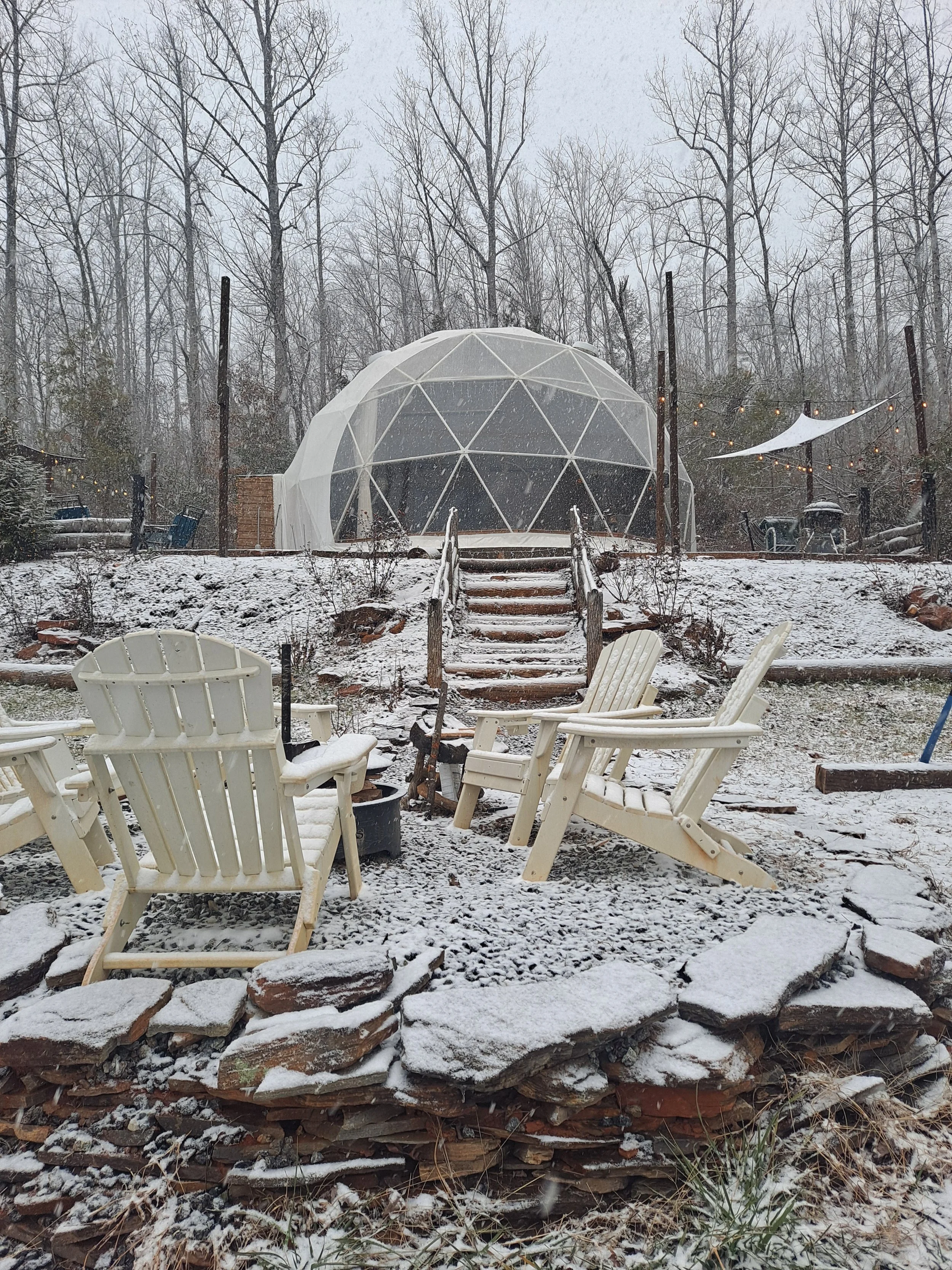 Snow-covered backyard with Adirondack chairs, stairs leading to a geodesic dome, and string lights.