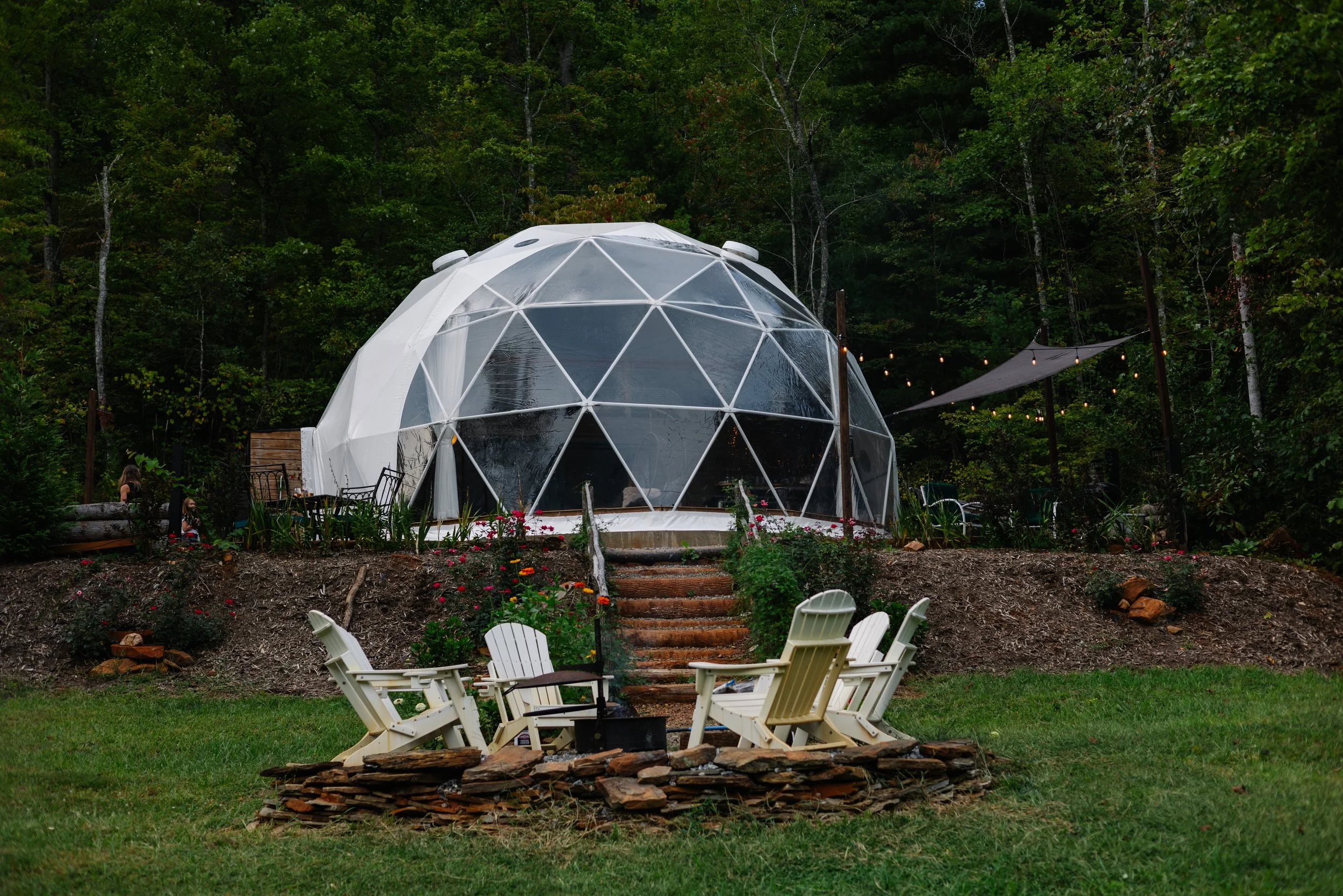 Geodesic dome on a hillside surrounded by greenery, with outdoor seating including Adirondack chairs and string lights.