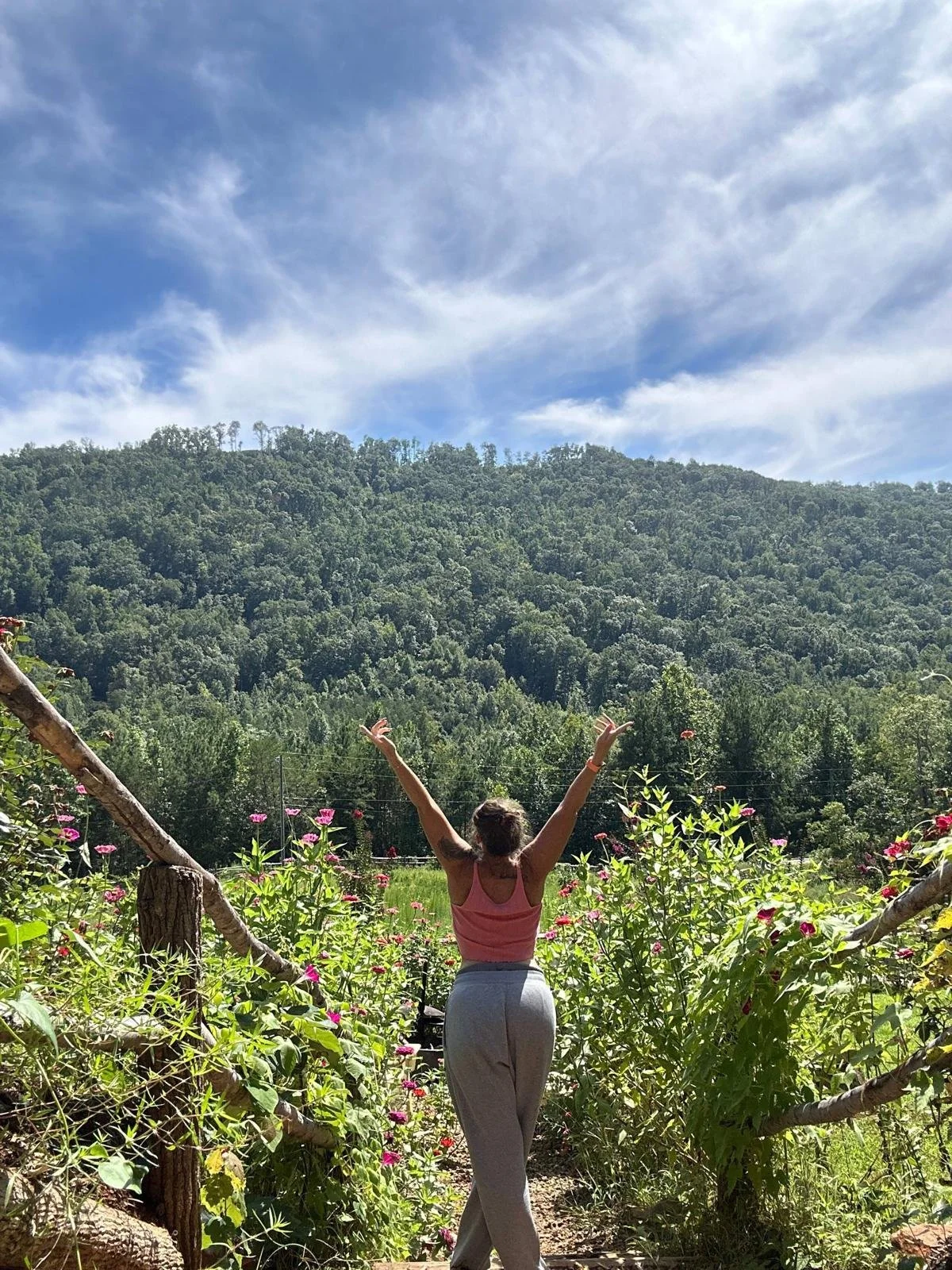 Person standing outside with arms raised, facing a green forested hill and blue sky with clouds.