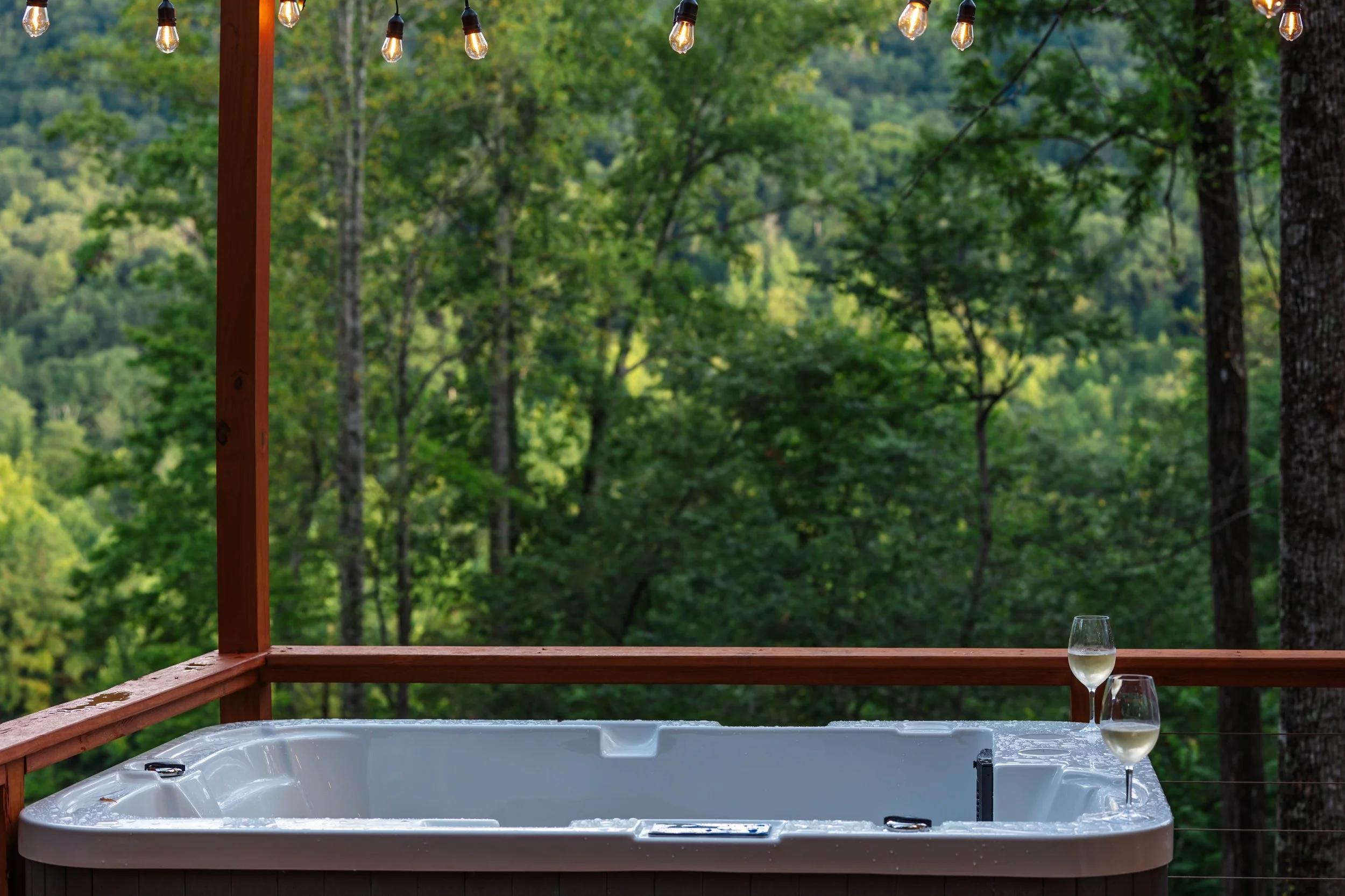 A hot tub with water, two glasses of white wine resting on the edge, set on a wooden deck with a view of a lush green forest and string lights hanging above.