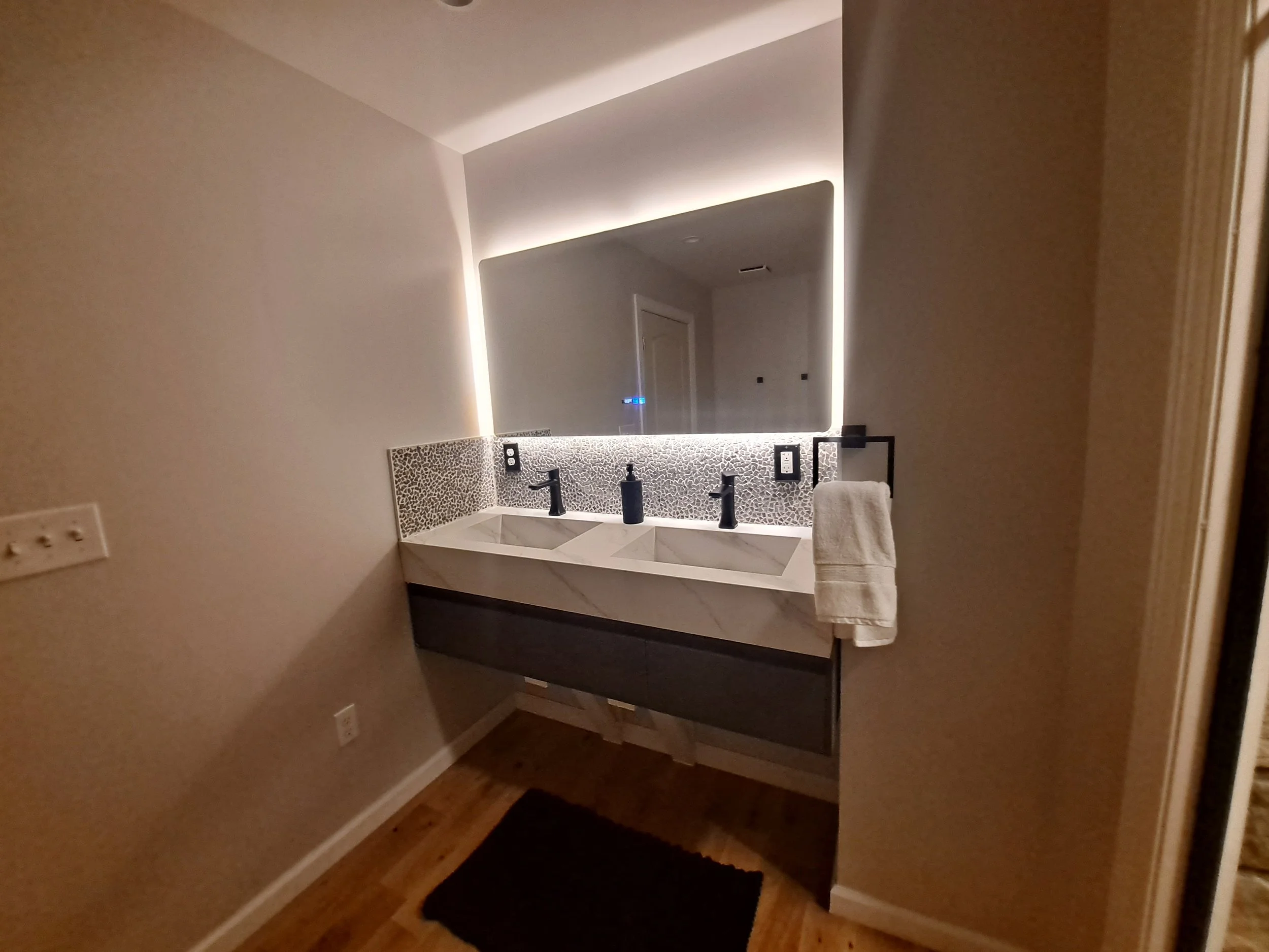 Modern bathroom vanity with a double sink, black faucets, a large backlit mirror, and black soap dispenser, with wooden flooring and a black rug in front.