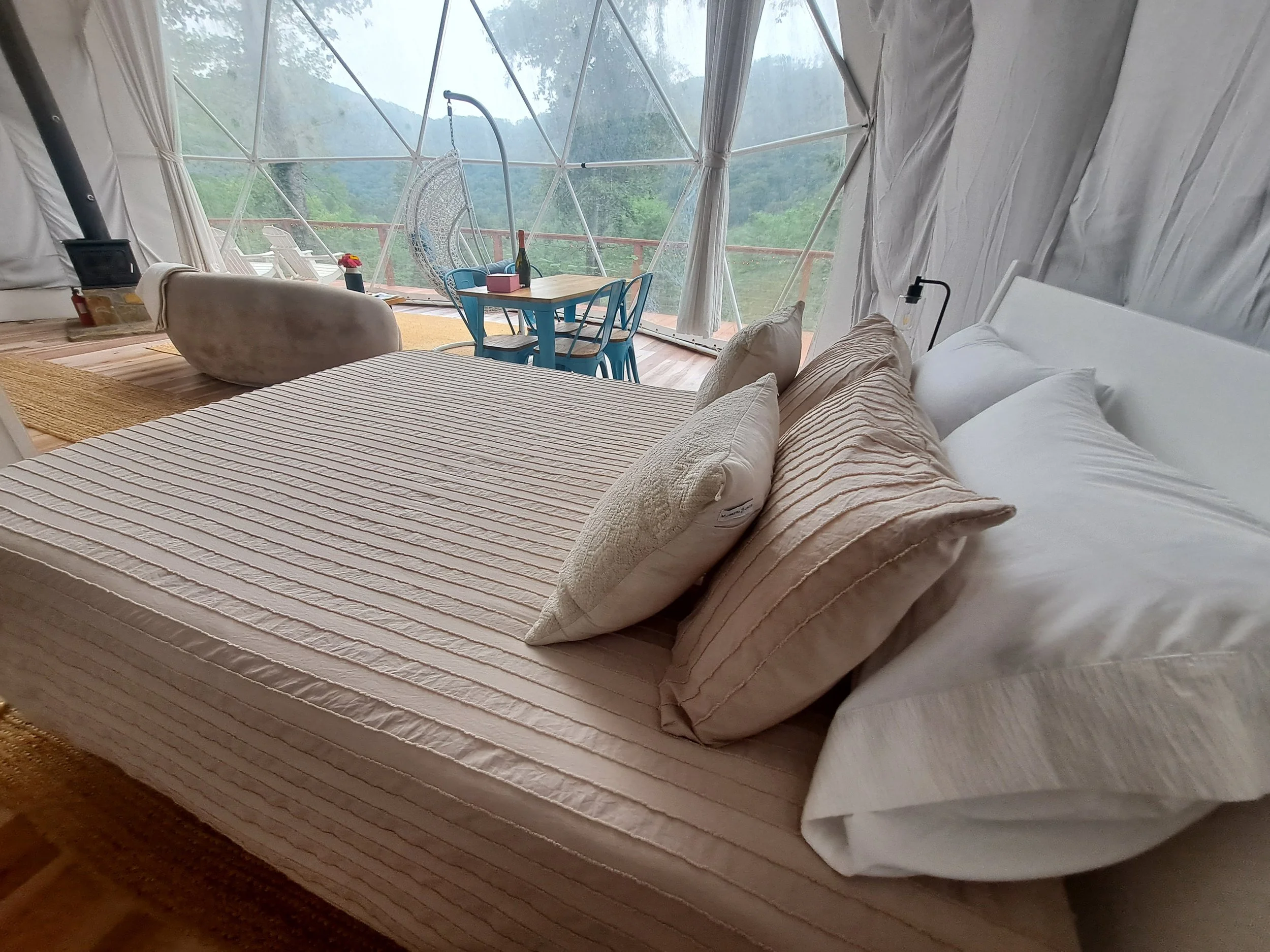 Inside a cozy dome tent with a bed with white and beige pillows, a small table with chairs, and a hanging chair near large geodesic window panels overlooking a lush green forest.