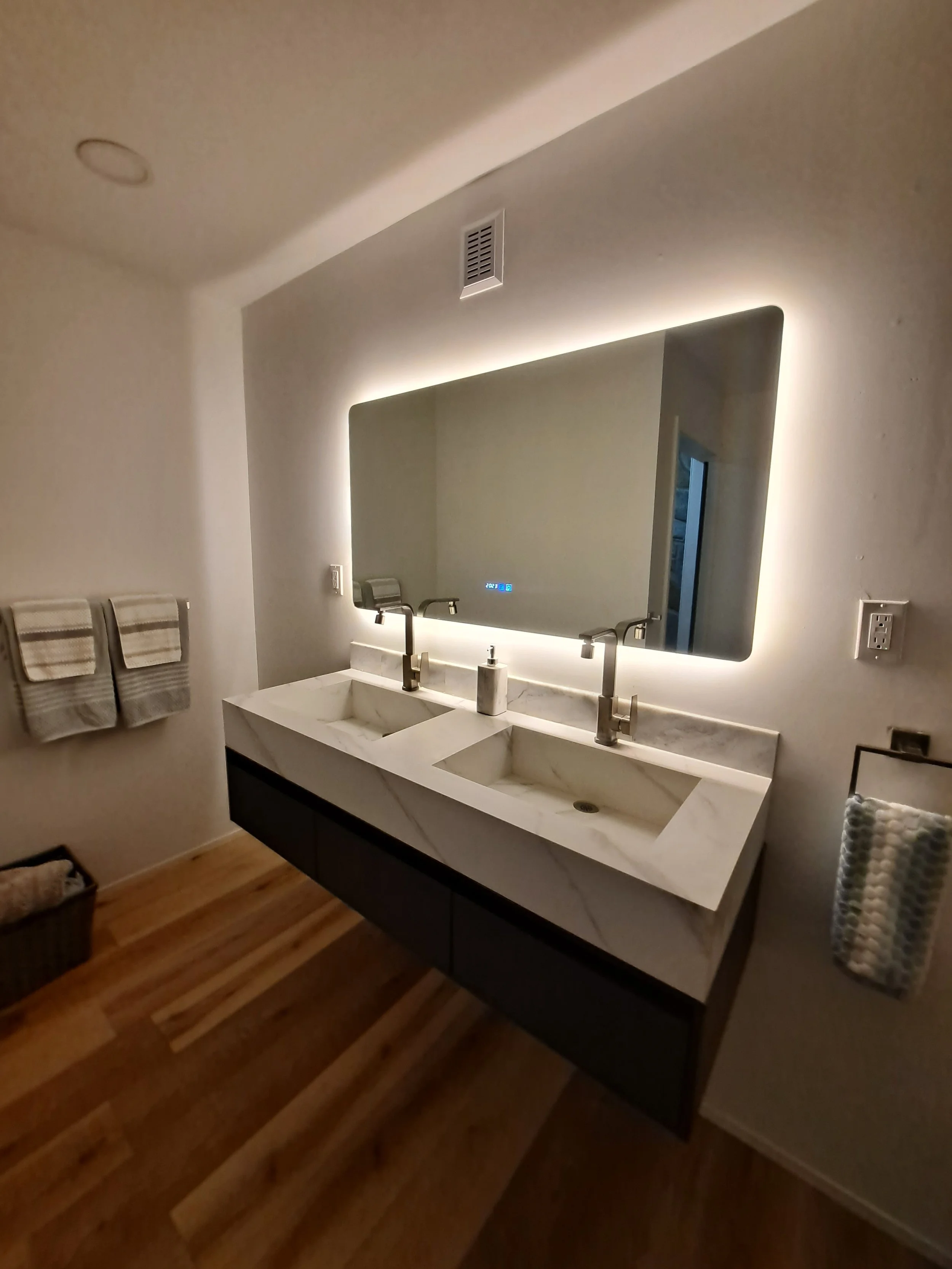 Modern bathroom with double marble sinks, backlit mirror, and wooden floor.