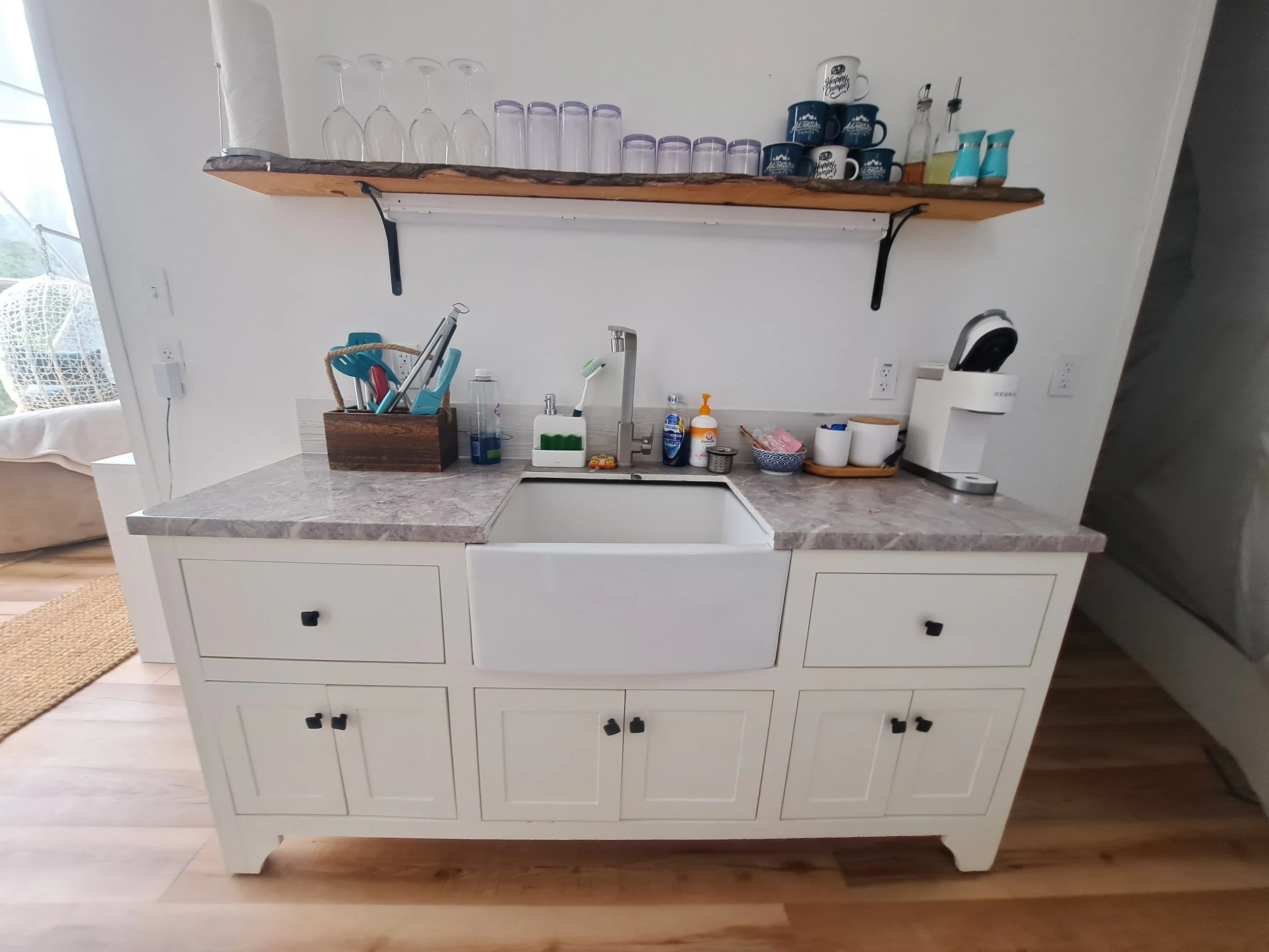Kitchen sink area with countertop and open cabinet with small drawers below. Items on the countertop include dish soap, a soap dispenser, a brush, a bowl, and a coffee machine. A wooden shelf above holds glassware, cups, and small bottles.