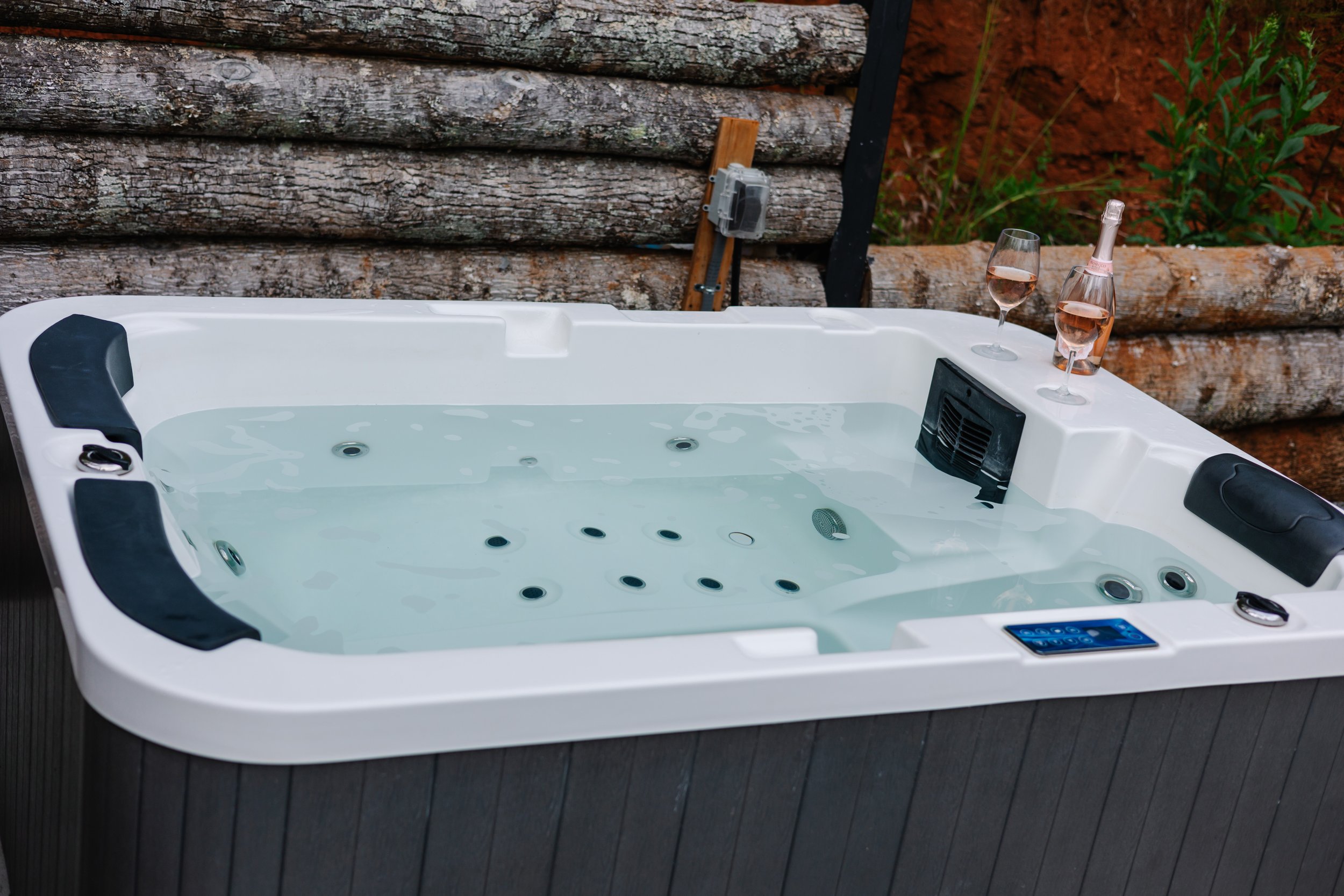 A white hot tub filled with water, placed outdoors with a log wall background. Two glasses of rosé wine and a pink glass bottle are on the hot tub's edge.