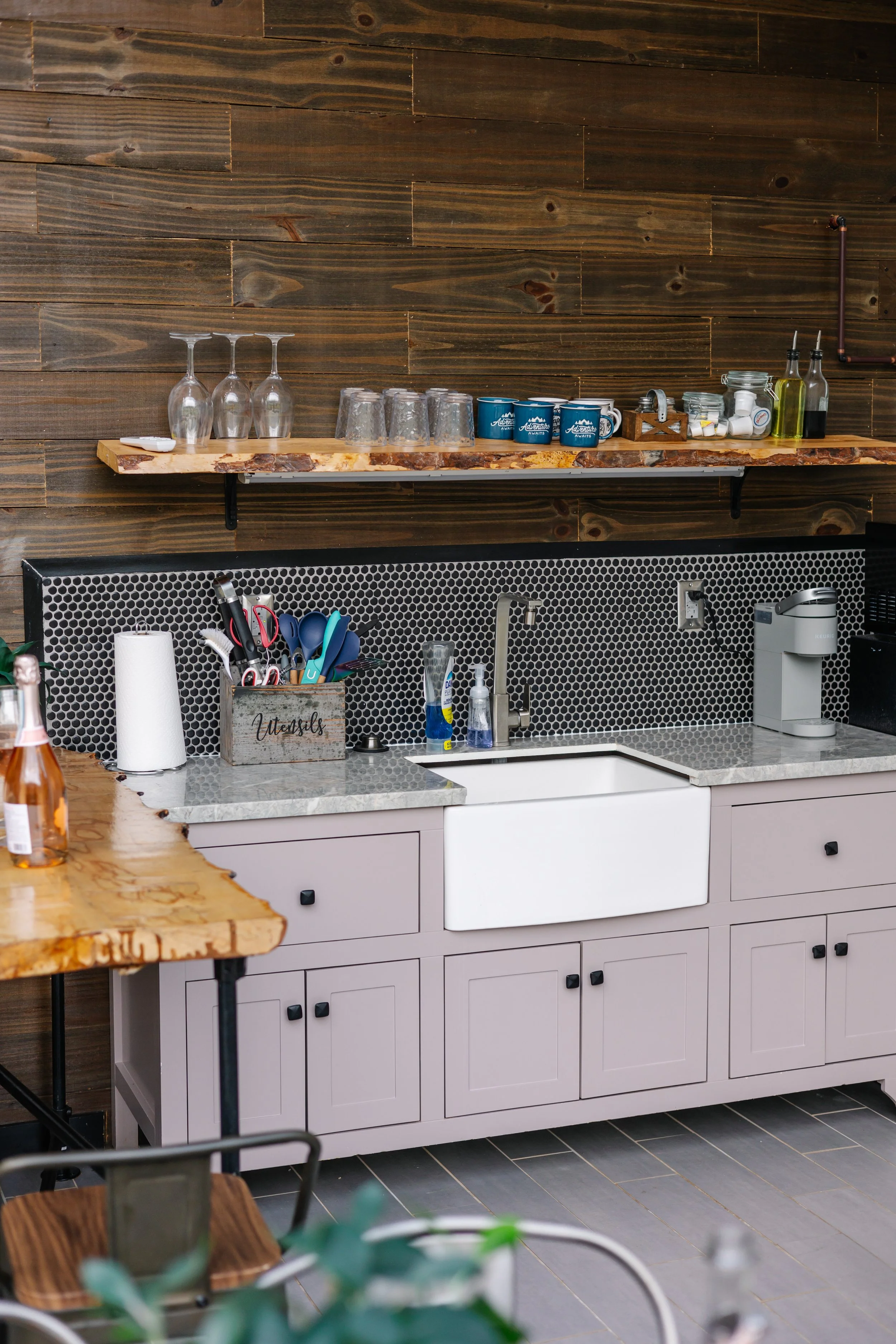 A modern kitchen with a light purple lower cabinet with black knobs, a white farmhouse sink, black honeycomb tile backsplash, and a dark wood wall. There is a wooden countertop on the left and a marble countertop above the sink with various kitchen u
